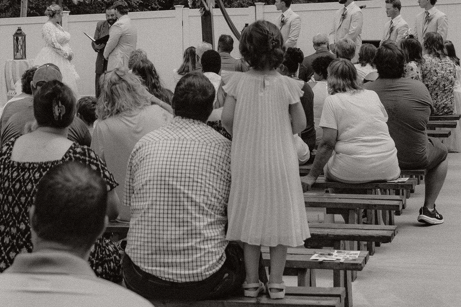 Black and white photo of wedding ceremony guests seated on wooden benches outdoors.