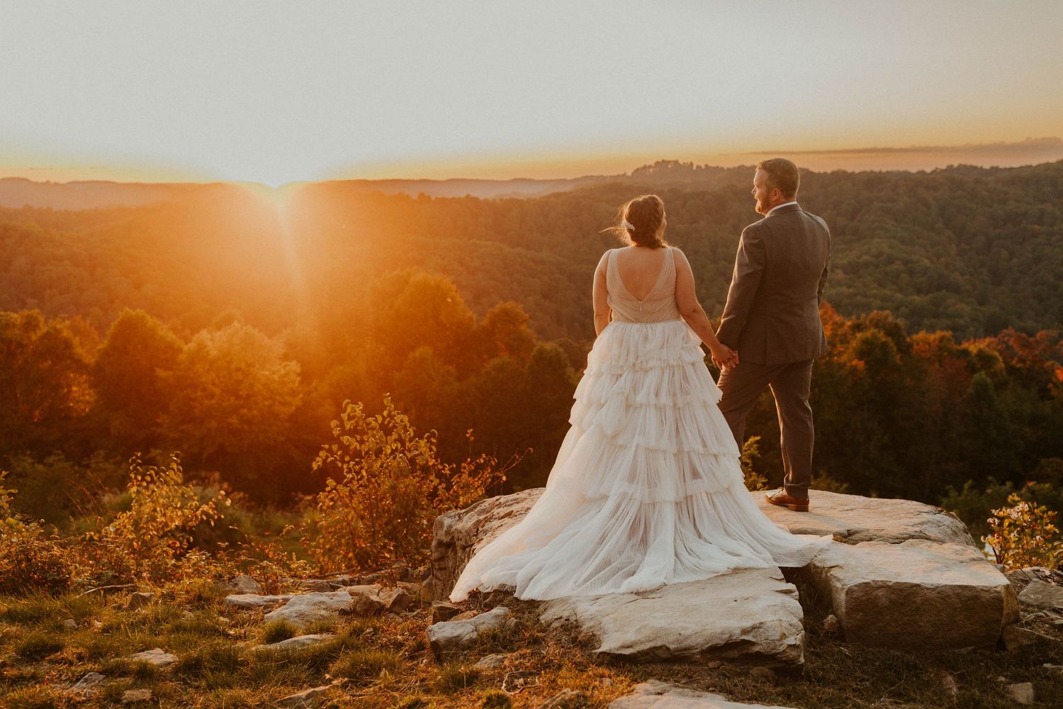 Couple stands on rocky overlook enjoying dramatic mountain sunset in wedding attire.