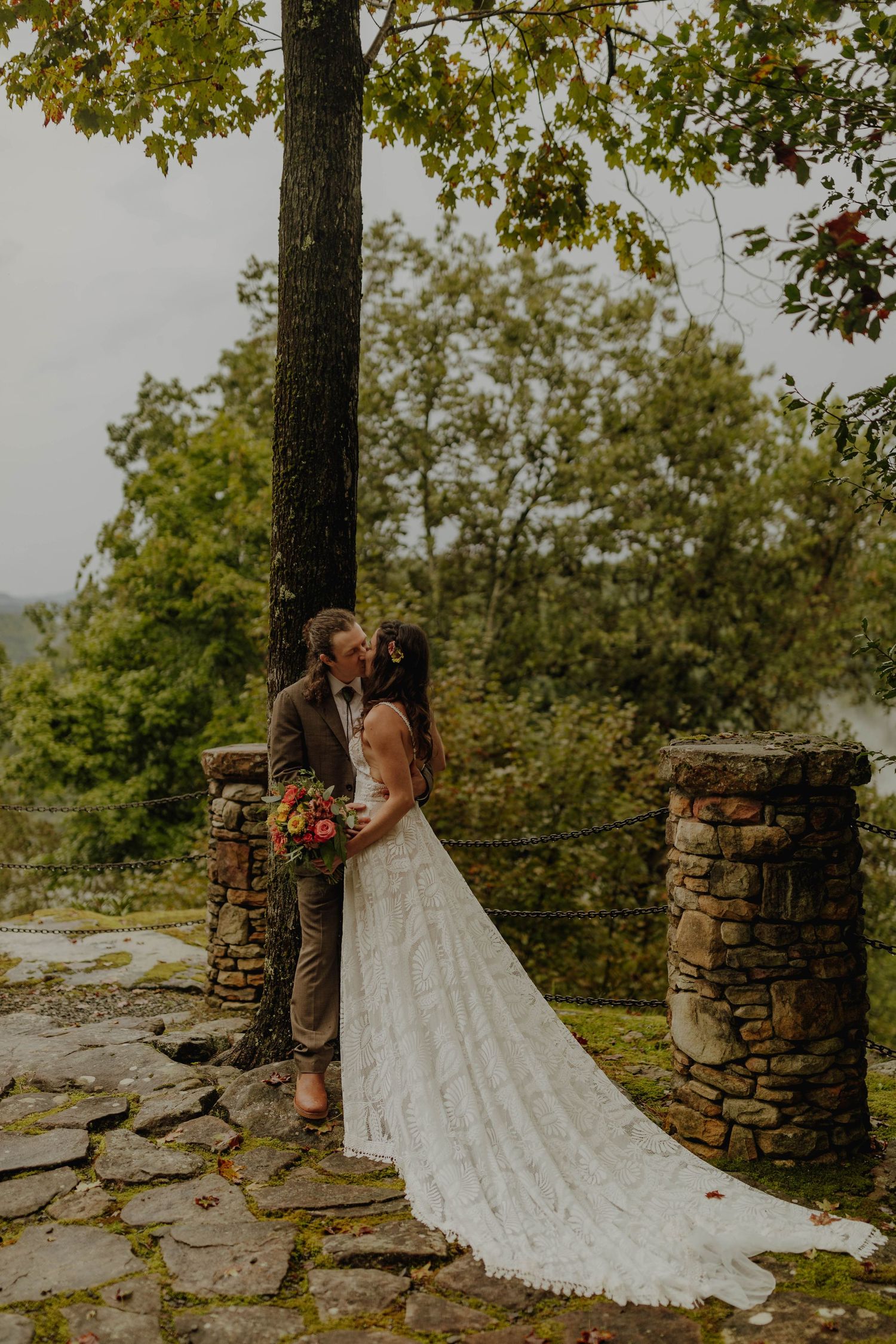 A wedding couple stands between stone pillars with a long lace train trailing behind.