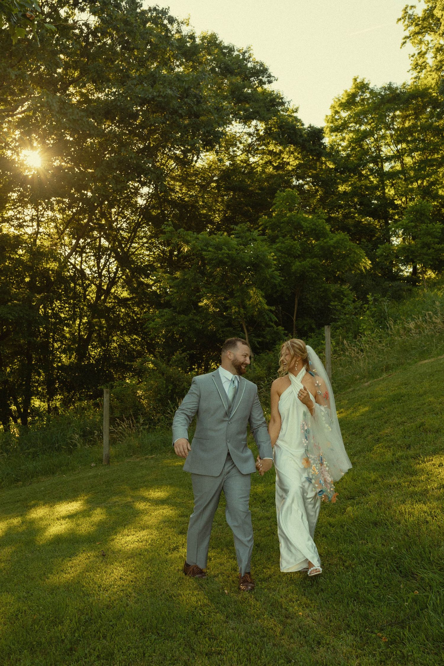 Bride and groom walk together on a grassy hillside during golden hour sunset surrounded by trees.