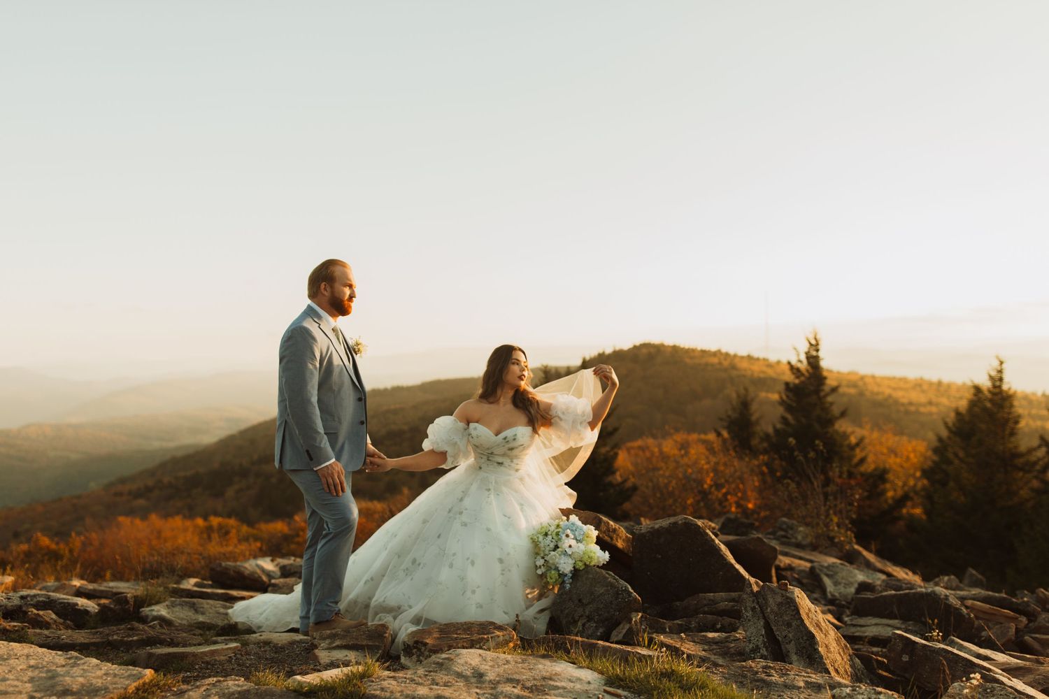 A couple in wedding attire stands atop a scenic mountain overlook at sunset.