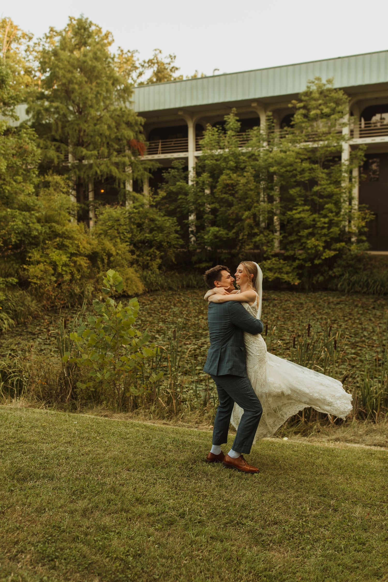 Newlyweds share intimate moment while dancing on grass with modern building in background.