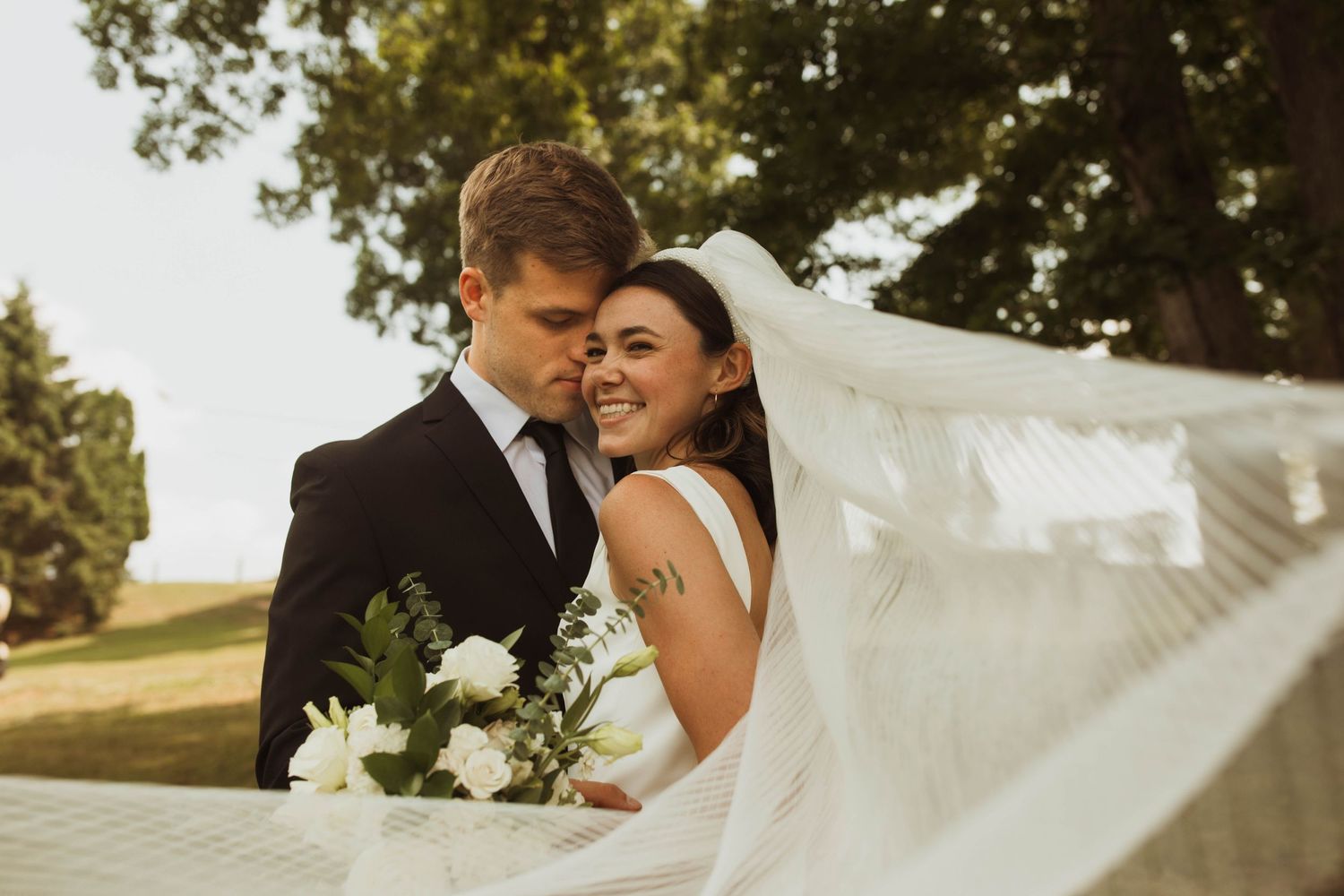 A joyful wedding moment captured with flowing veil and white flowers in warm sunlight.