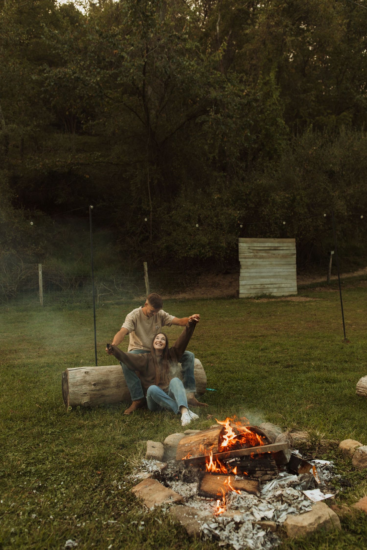 Cozy evening scene with couple sitting by rustic campfire on wooden log in grassy field.