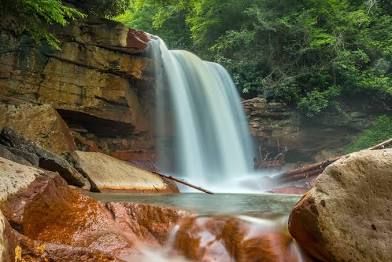 A majestic waterfall cascades over red rocks in a lush forest setting.