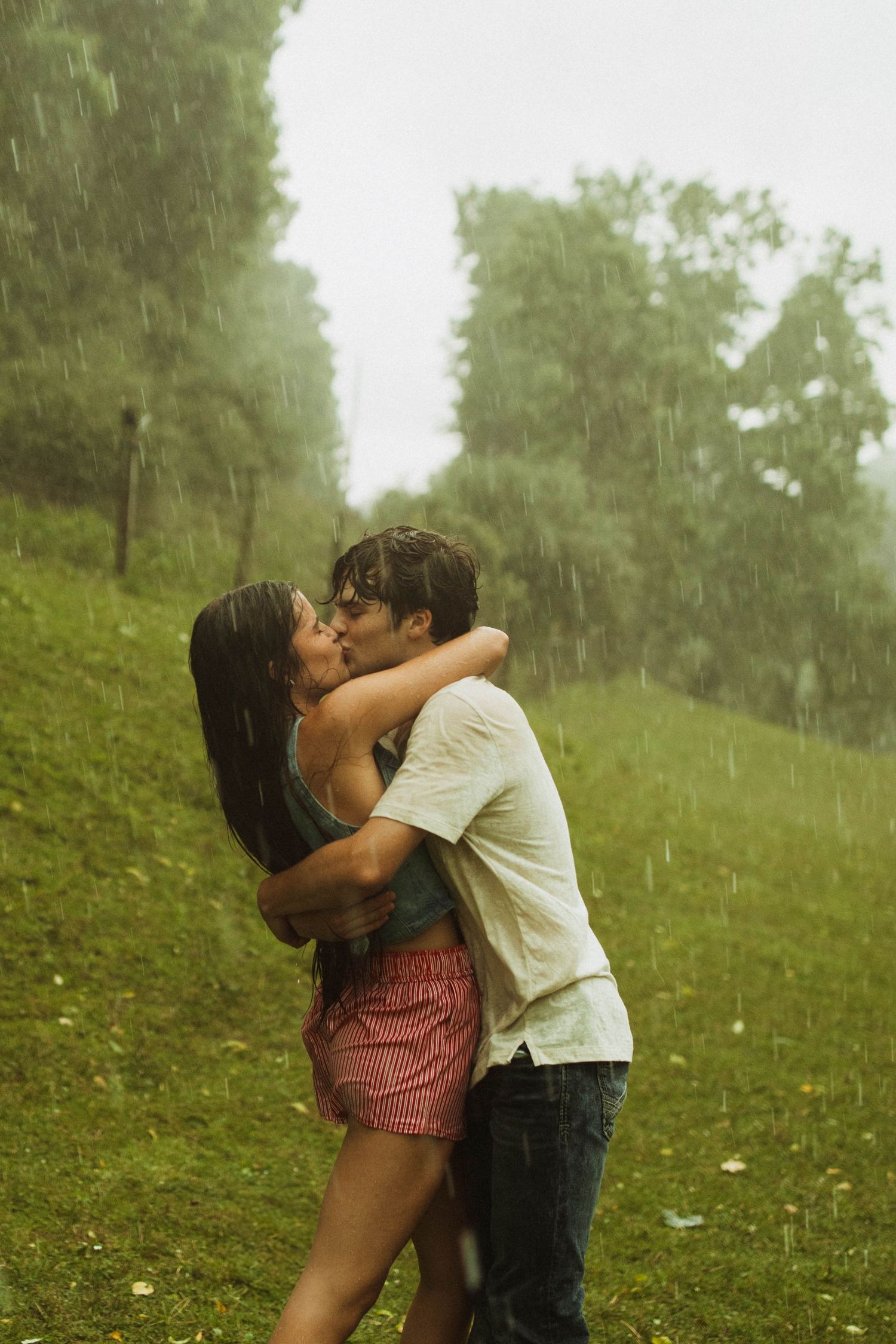 A romantic couple embraces in the rain on a lush green hillside.