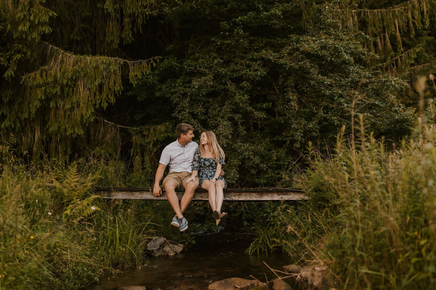 A couple sits together on a rustic wooden bridge surrounded by lush greenery in a romantic forest setting.