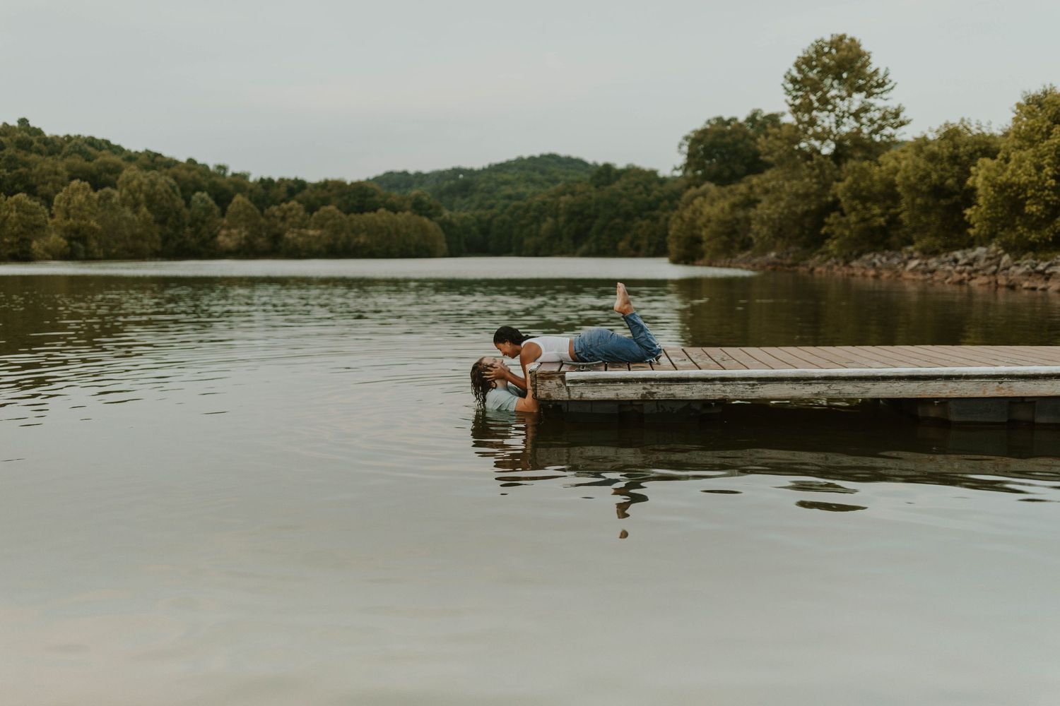 A couple sits peacefully on a wooden dock overlooking a serene lake surrounded by green hills.