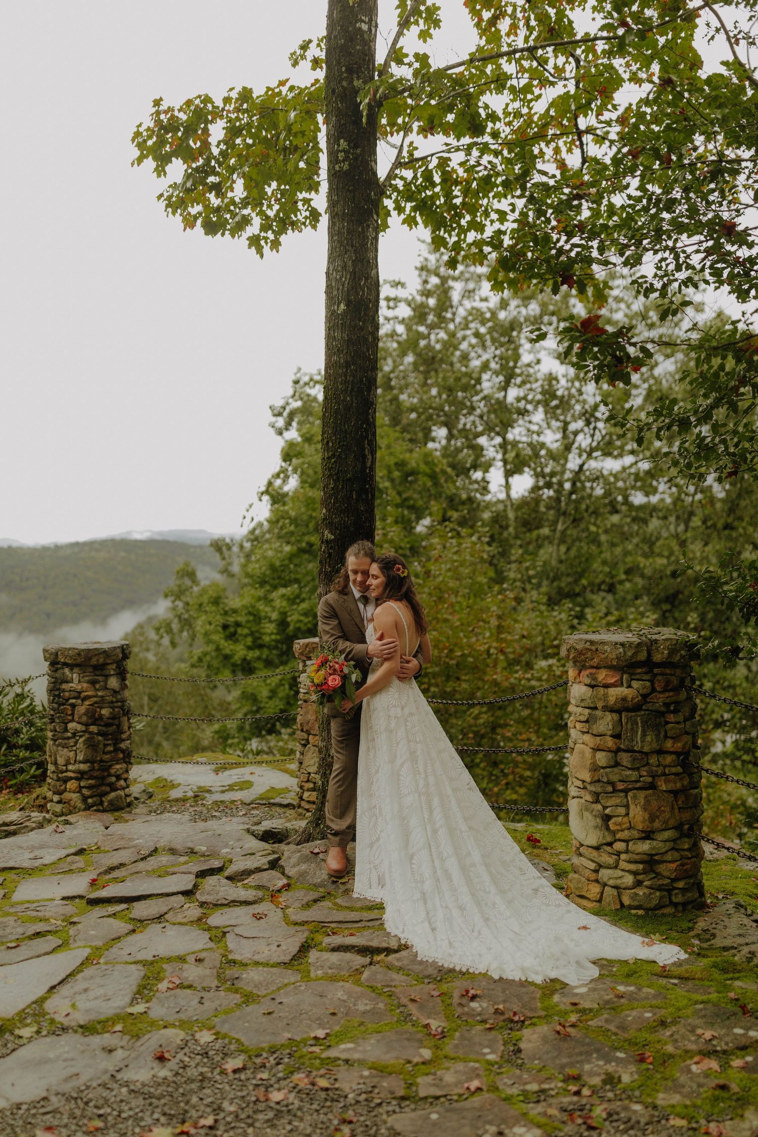 A couple embraces by stone pillars on a scenic mountaintop overlook during their wedding day.
