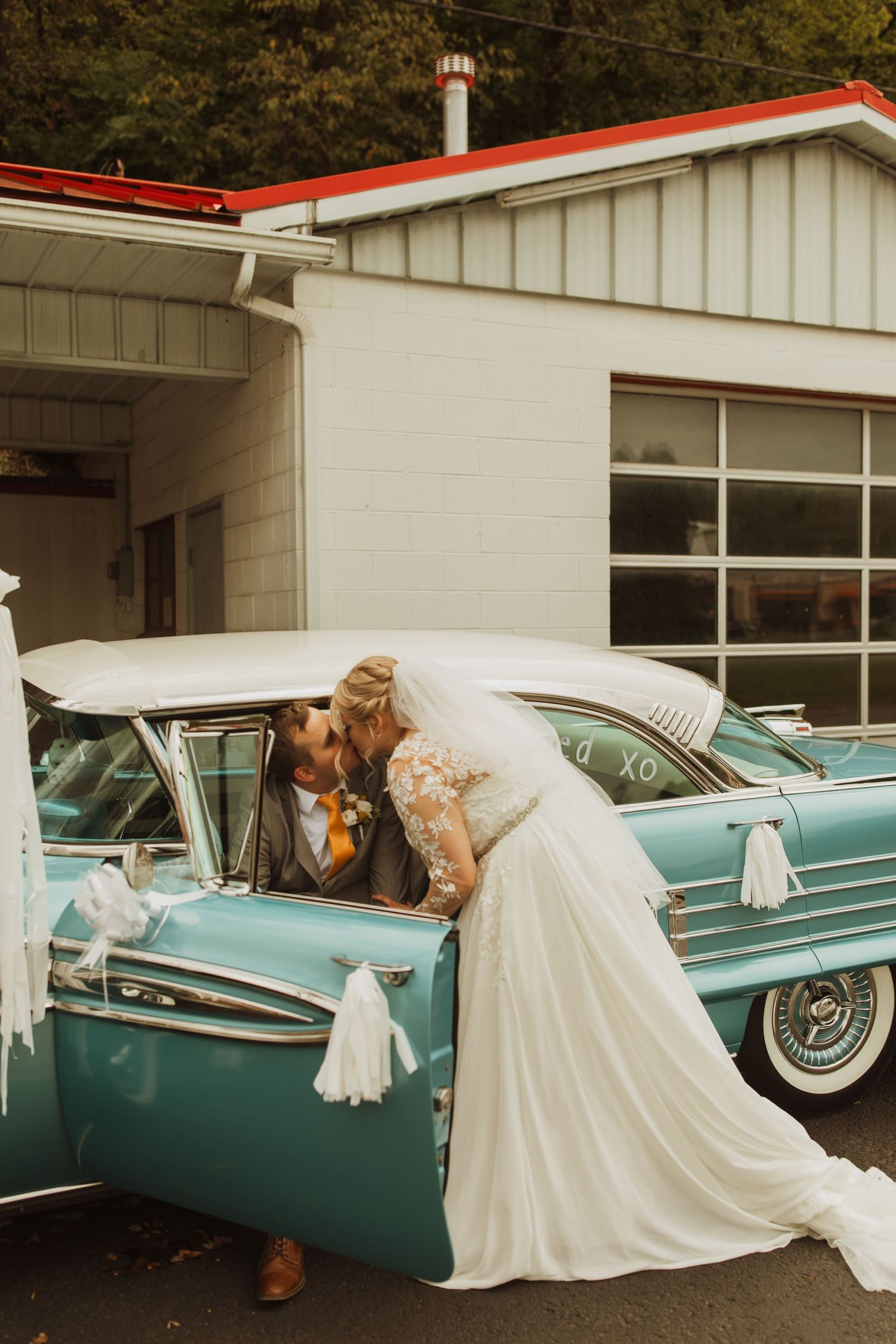 A couple poses with a classic turquoise 1950s car outside a vintage-style building.