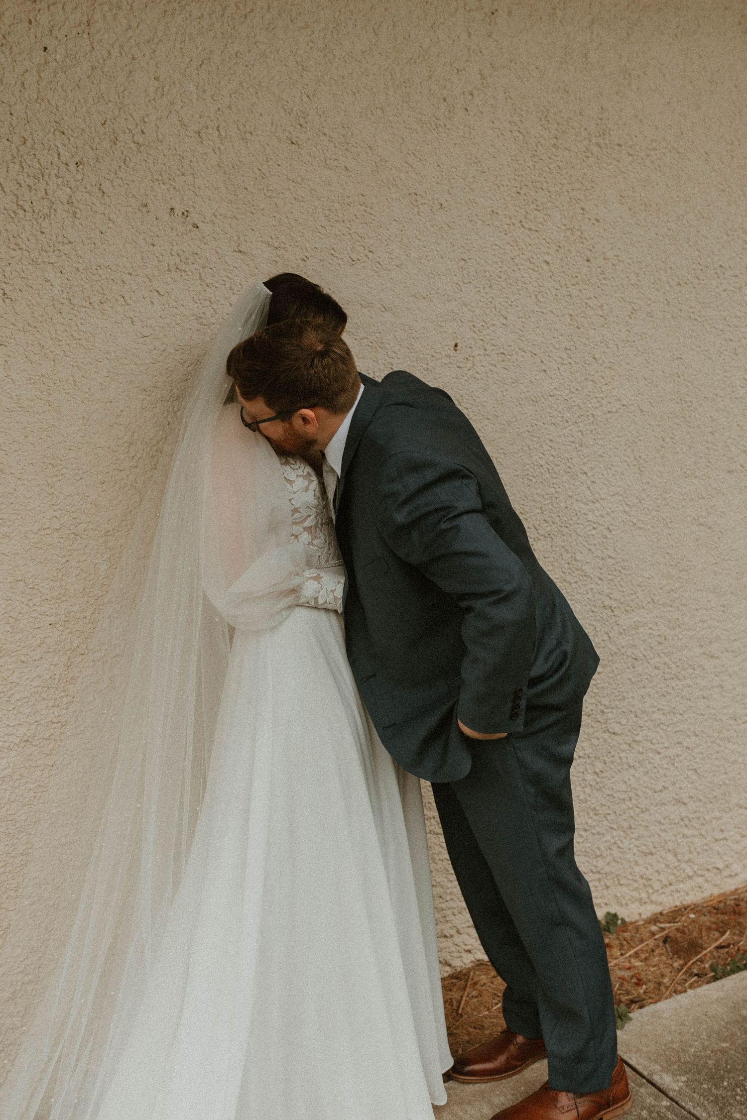 A tender moment between newlyweds standing against a beige wall, with the bride in a flowing white dress and groom in dark suit.