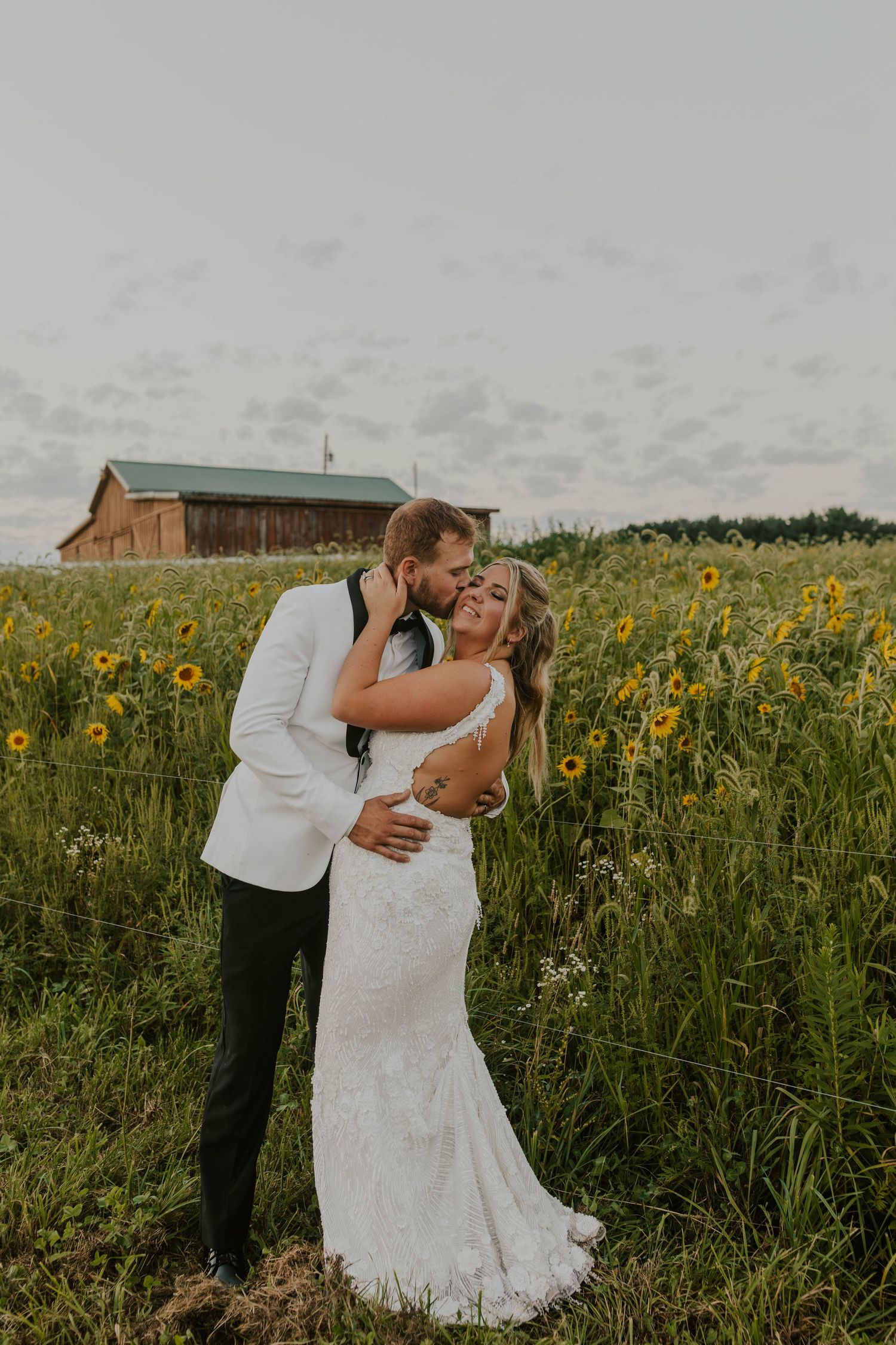 Couple embracing in a sunflower field with rustic barn in background.