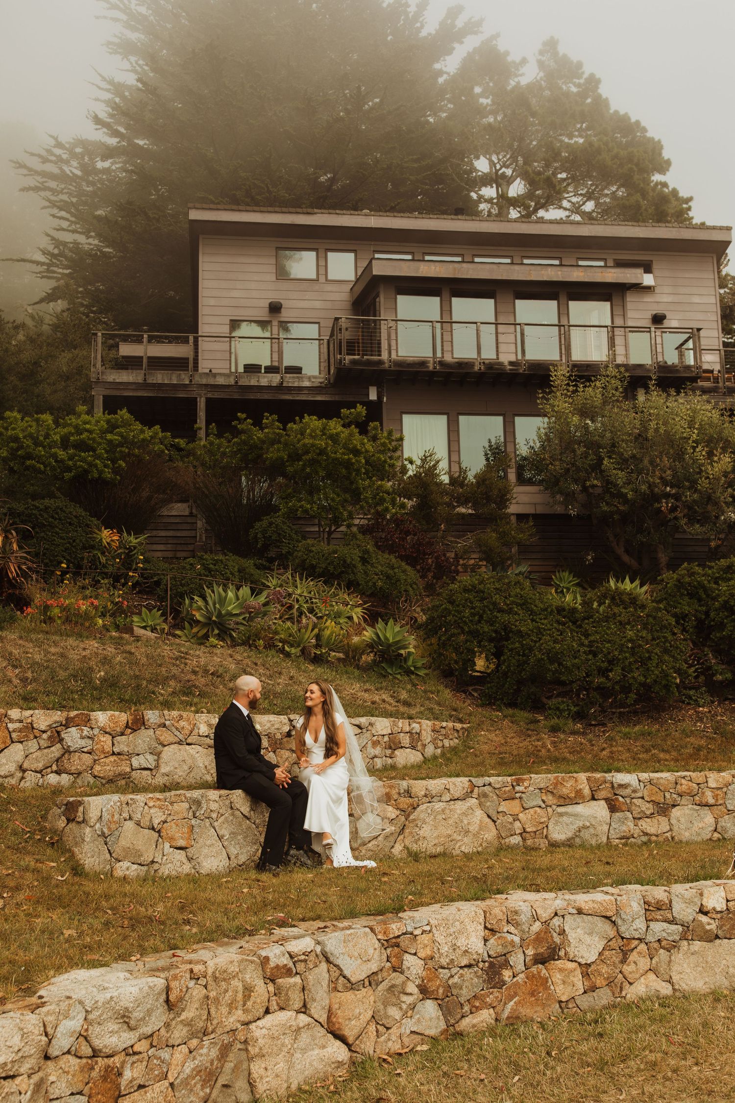 A couple sits on stone terraces beneath a modern house on a foggy hillside.