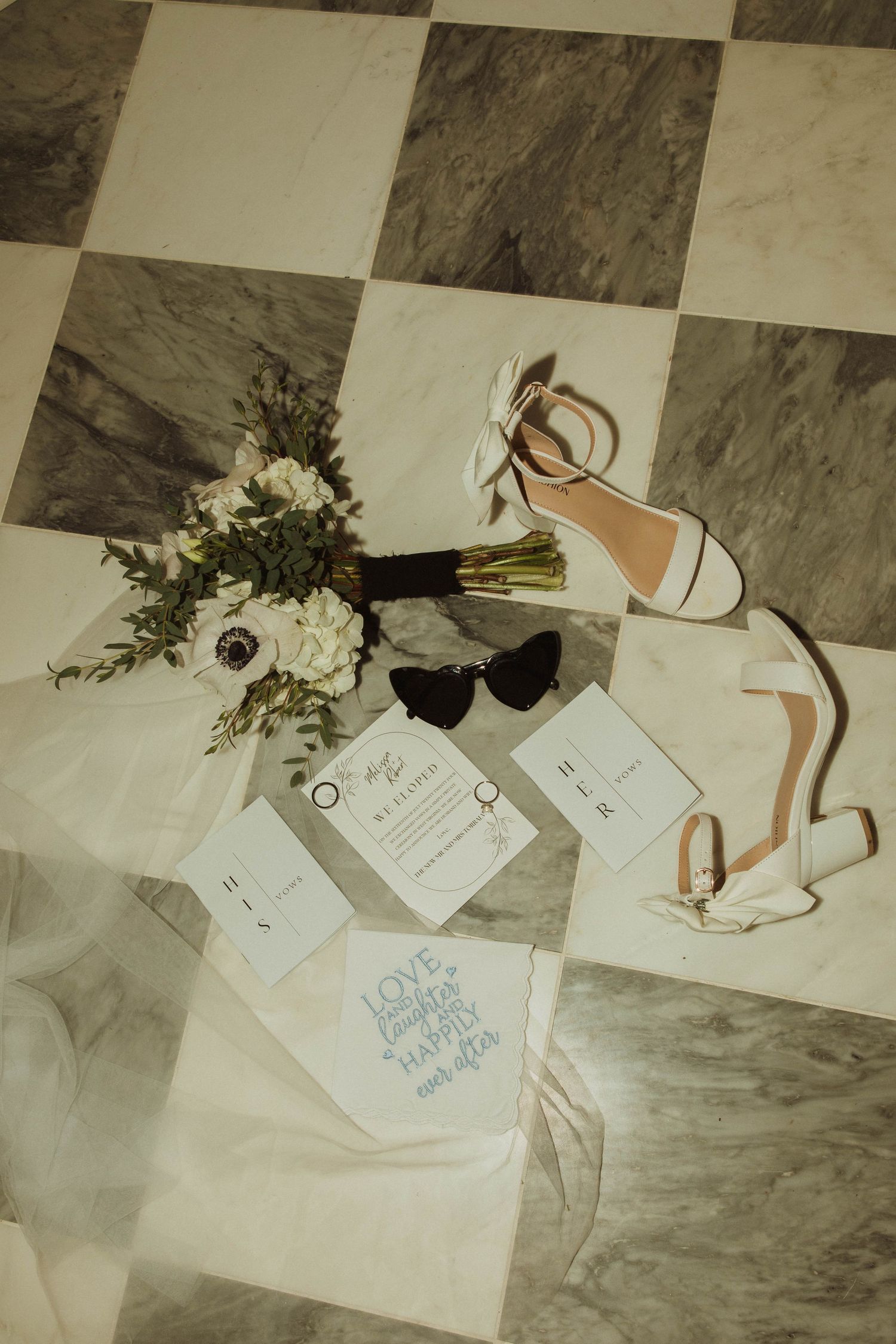 Wedding details flatlay with invitations, white flowers, and strappy sandals on checkerboard floor.