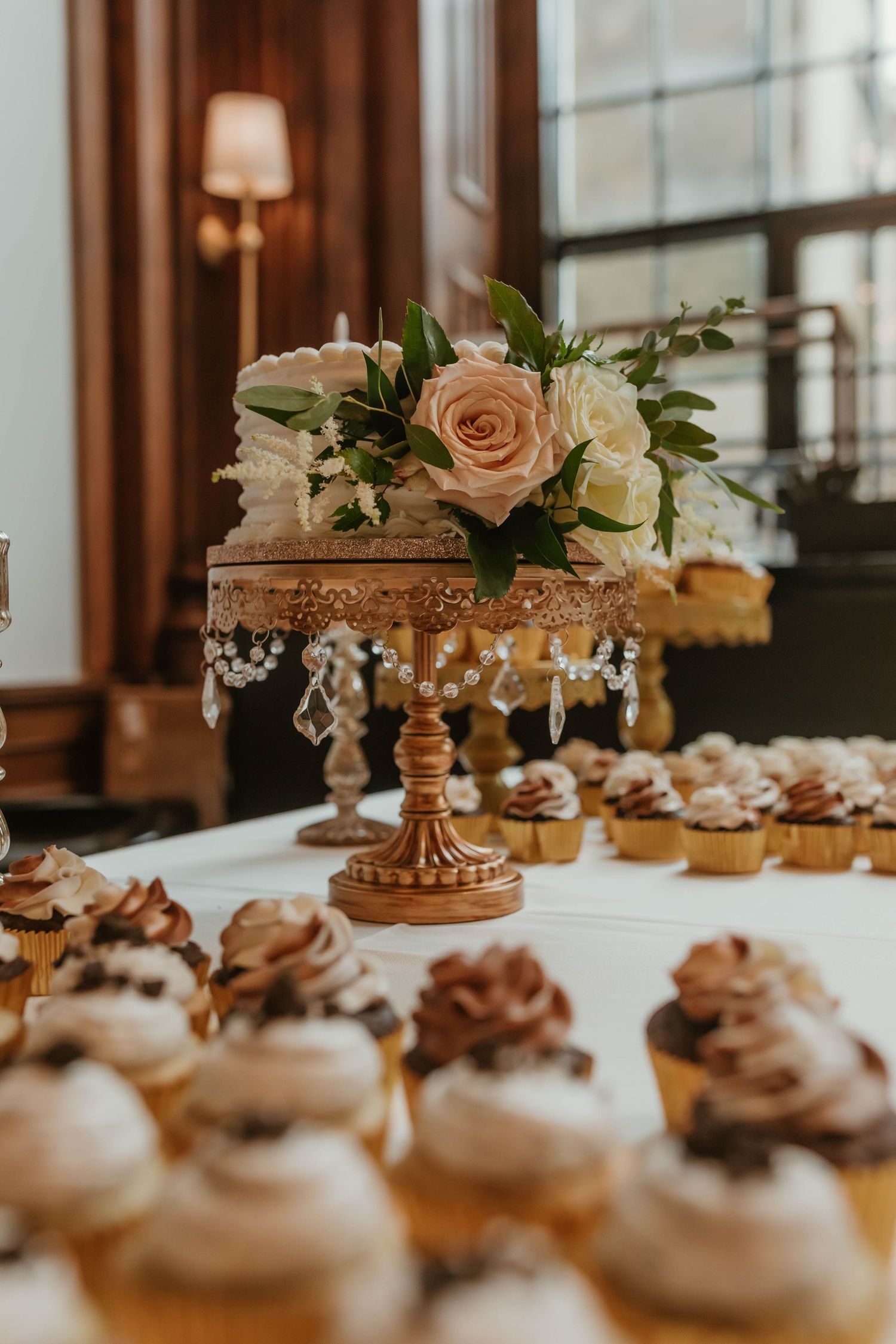 Wedding dessert table with cupcakes and floral centerpiece on ornate gold stand.