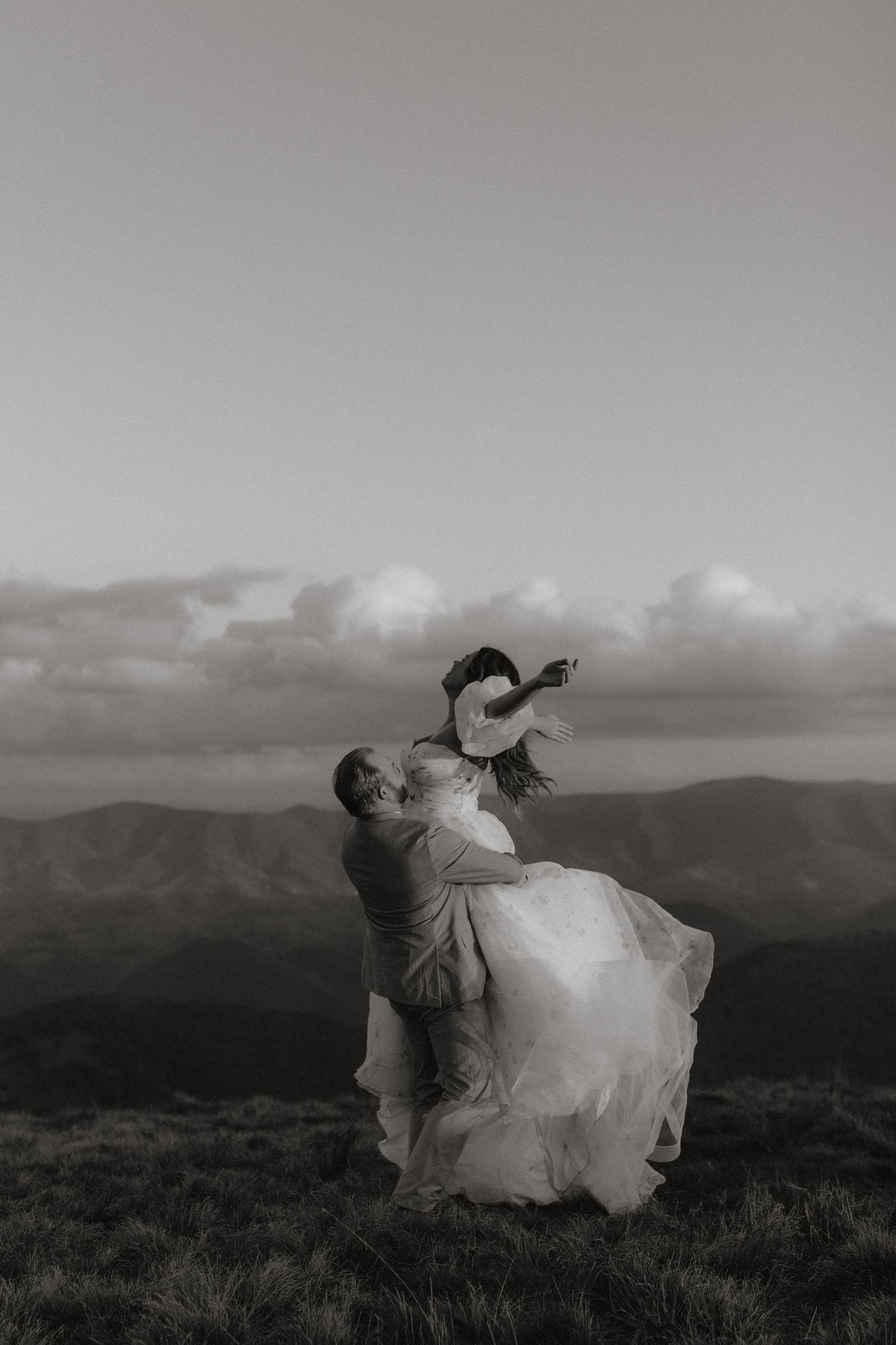 A romantic wedding couple dances atop a mountain with dramatic cloudy skies in black and white.