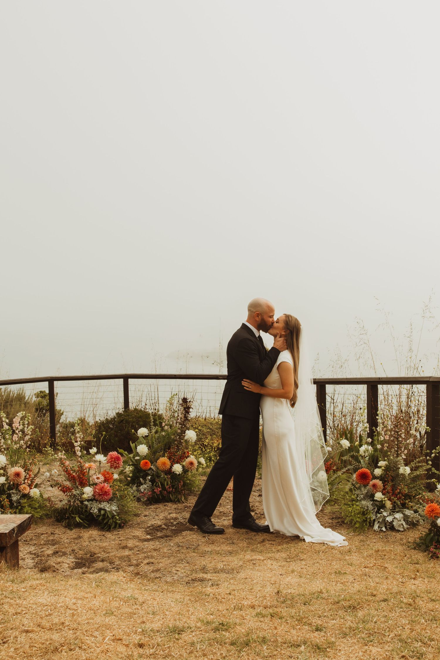 A wedding couple embraces on a foggy hillside with colorful floral arrangements decorating the ground.