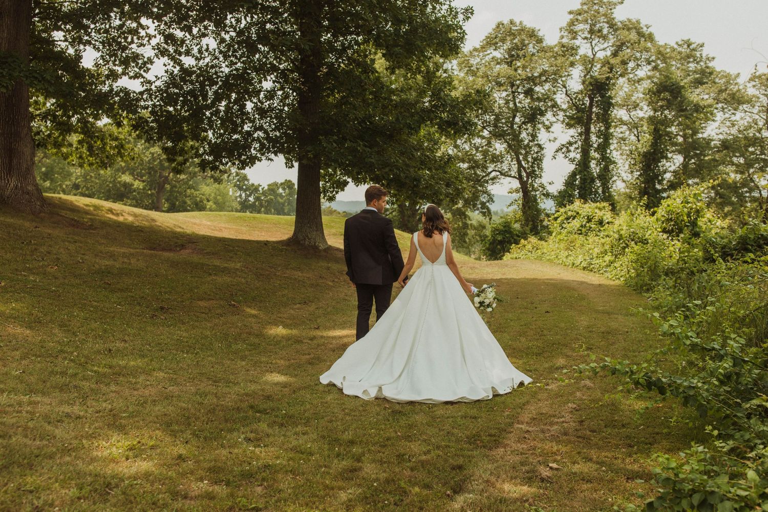 A classic full-length wedding portrait showing a couple walking on a hill under mature trees.