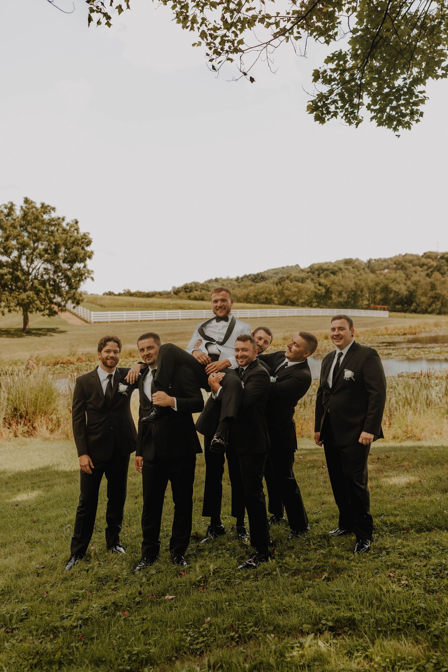 Groomsmen in black suits pose playfully on grassy hill with lake in background.