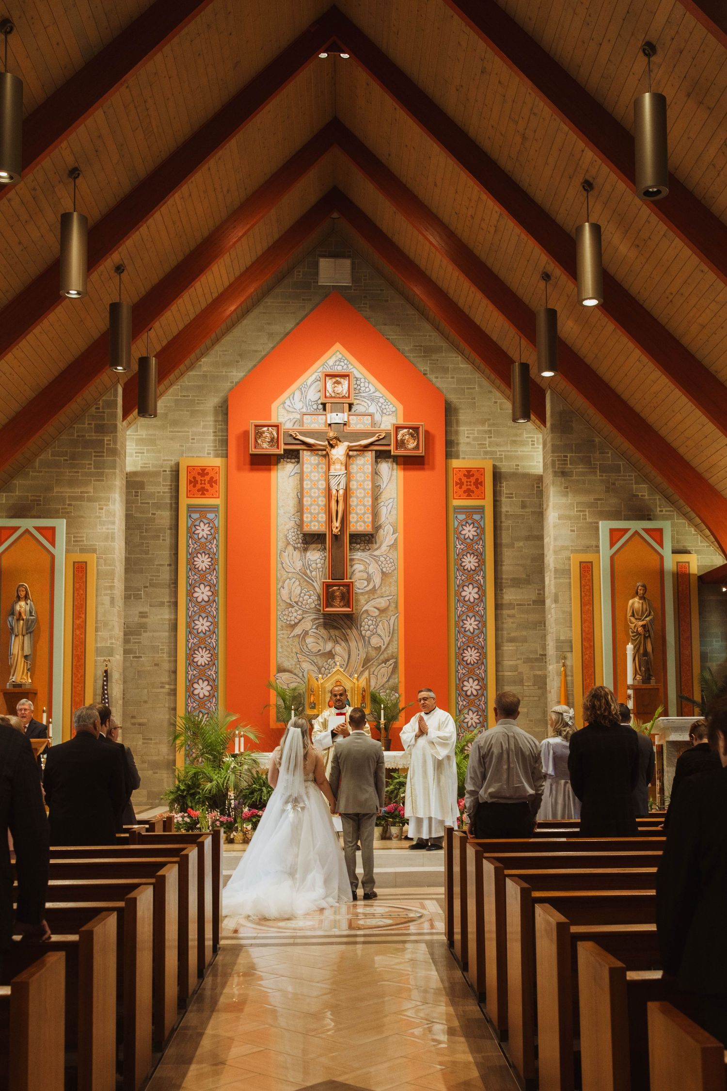 Interior of a church during wedding ceremony with orange cross altar and wooden beams overhead.