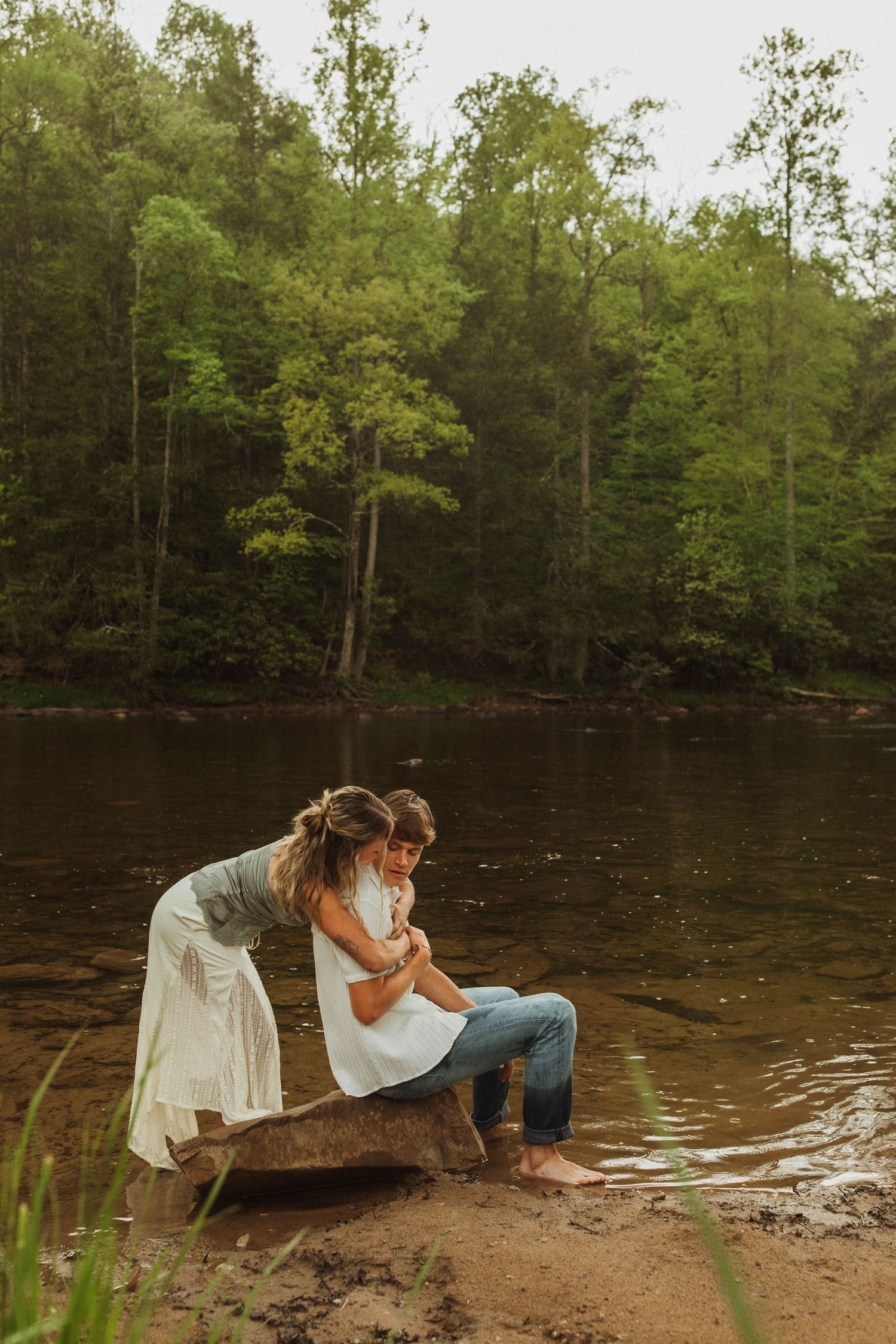 A couple shares an intimate moment while sitting on rocks by a peaceful forest lake.