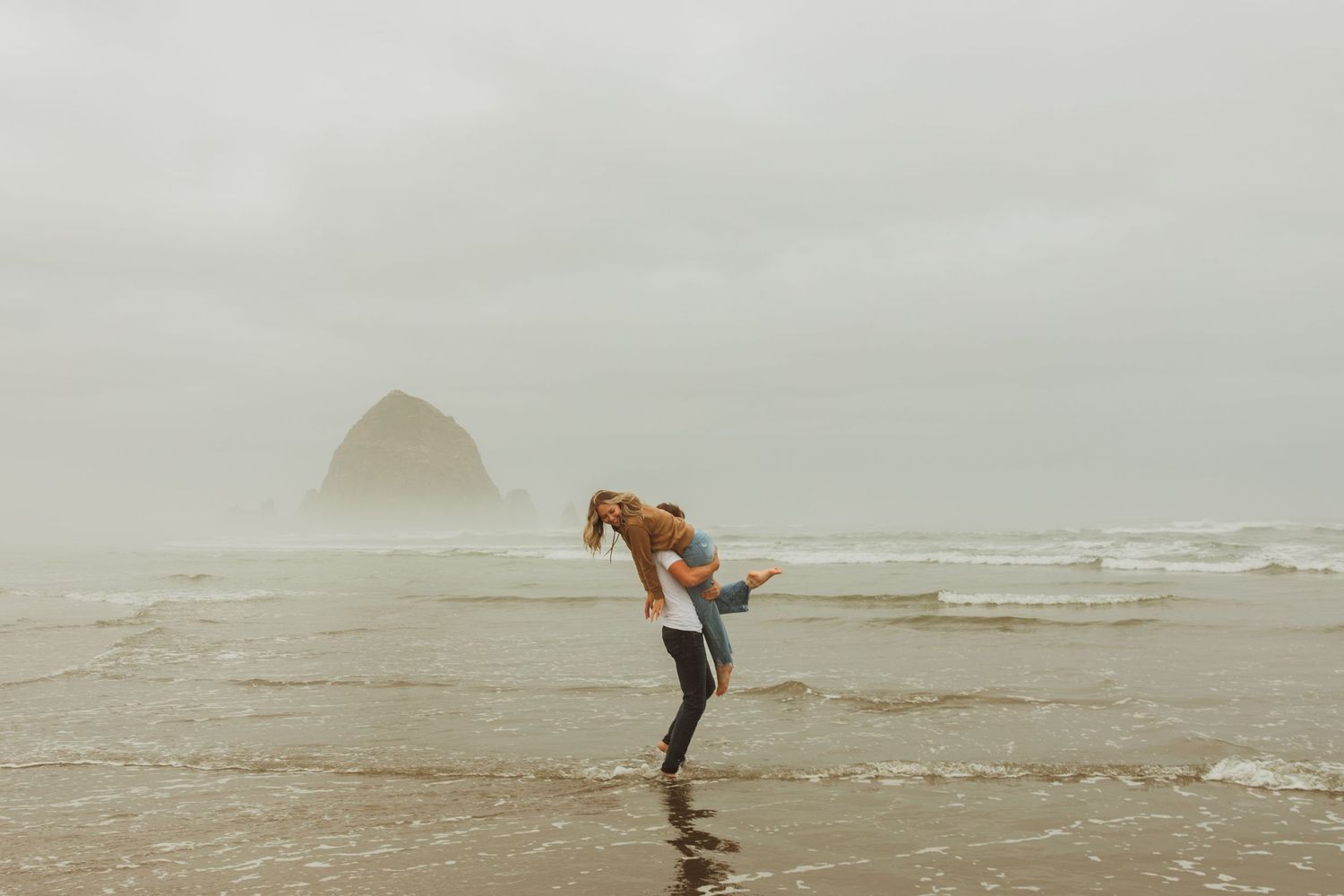 Playful scene on a misty beach with figures enjoying the waves near a distinctive rock formation in the distance.