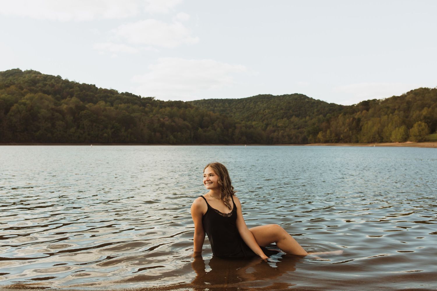 Peaceful lake scene with swimmer in dark swimsuit enjoying calm waters surrounded by forested hills.