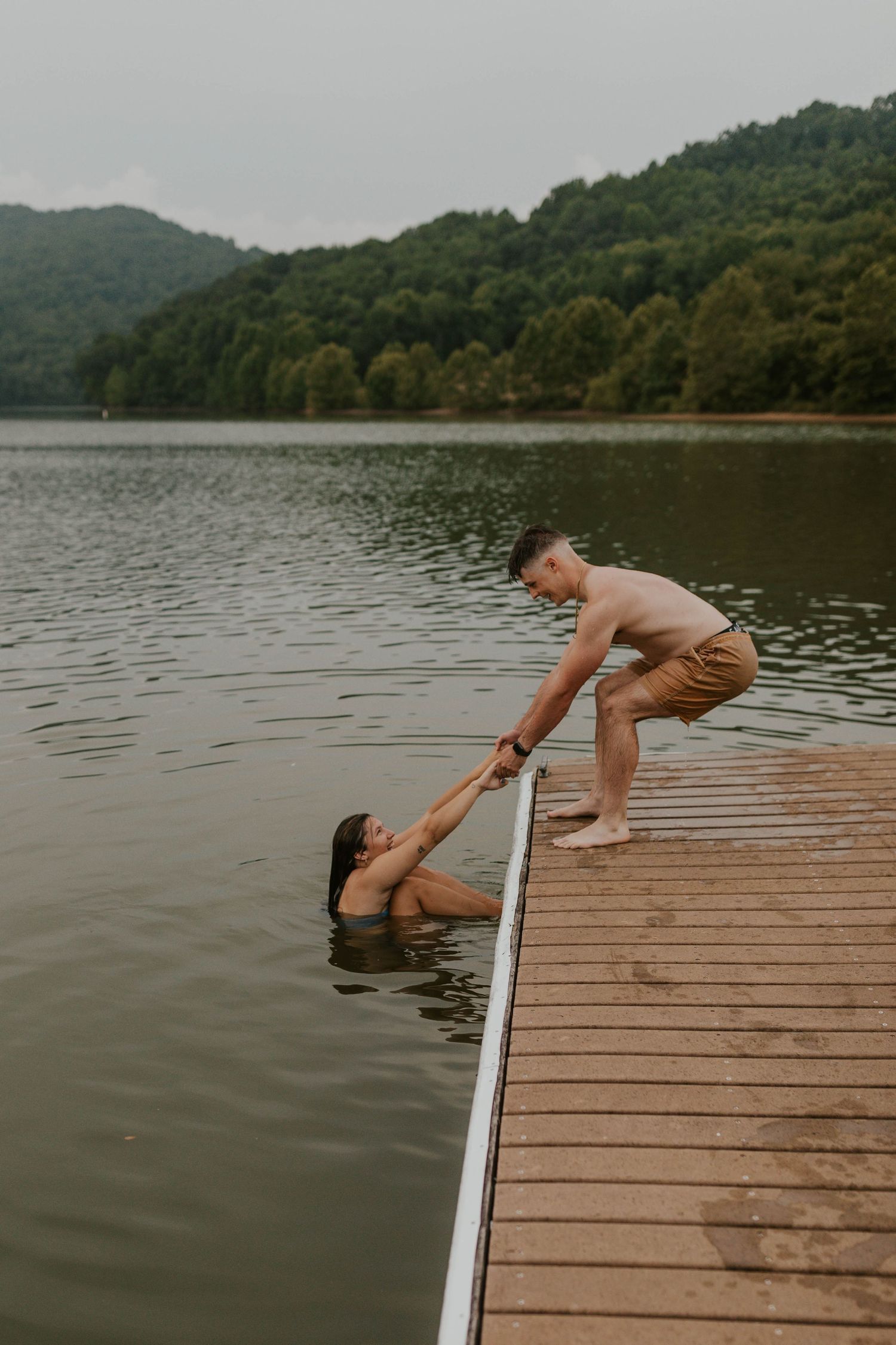 Person helping someone out of the water on a wooden dock at a lake.