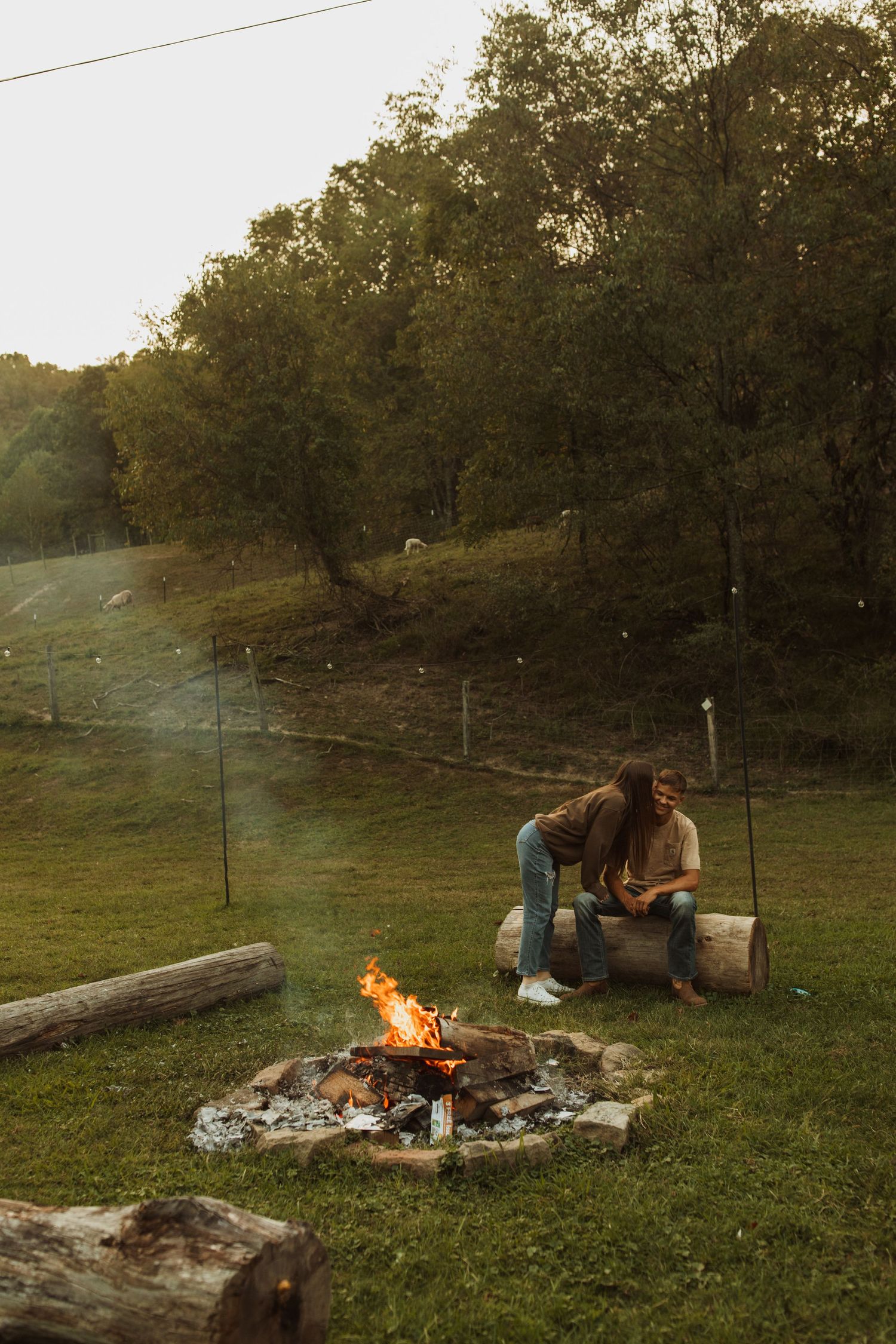 Intimate moment shared on log bench next to glowing campfire at dusk.