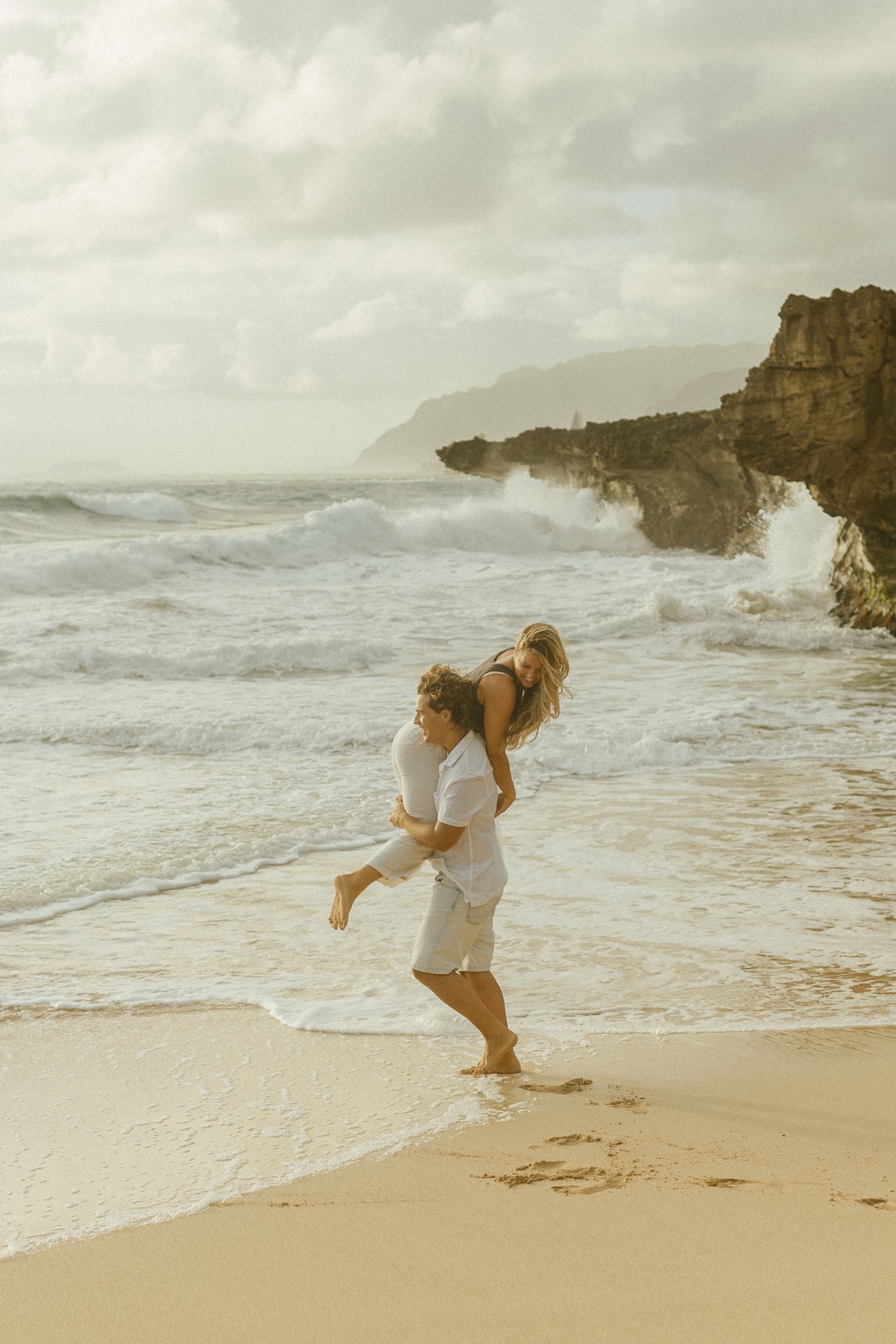 Romantic beach scene with couple playing in the waves near dramatic coastal cliffs.