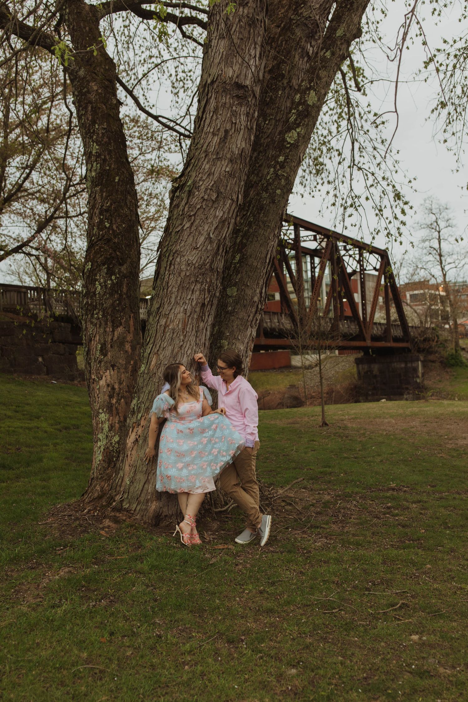 Romantic scene under large tree with vintage railway bridge in background.