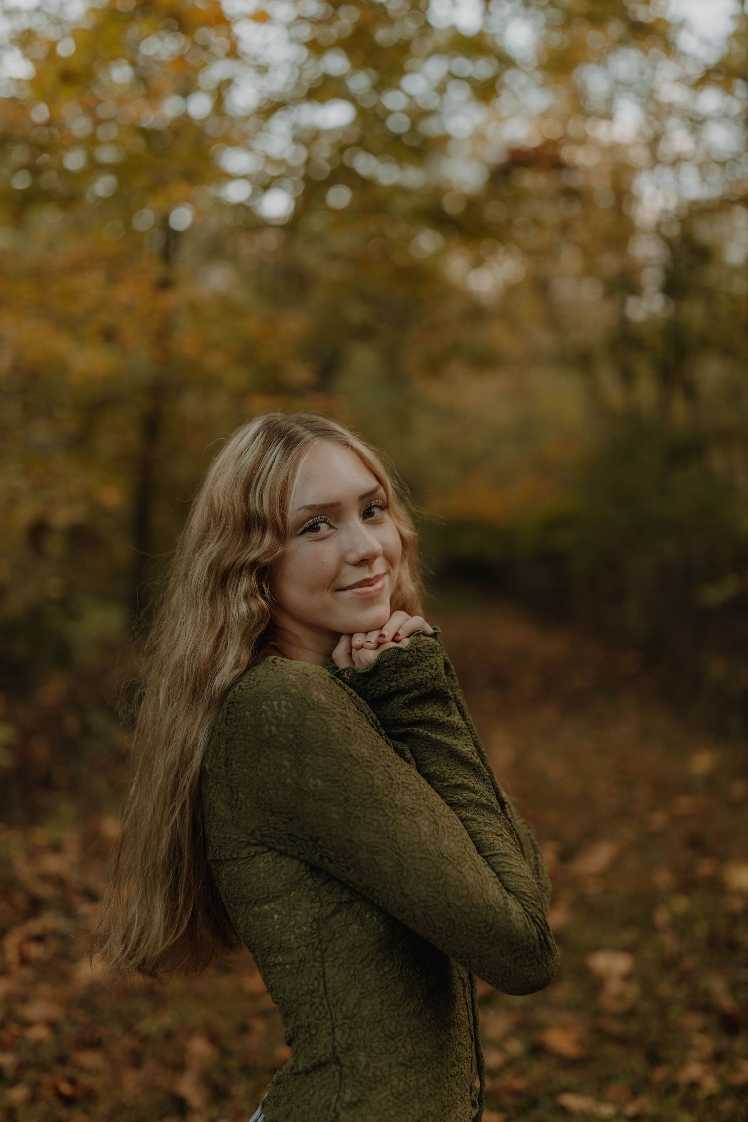 A person wearing a green sweater poses against an autumn forest backdrop in warm moody lighting.