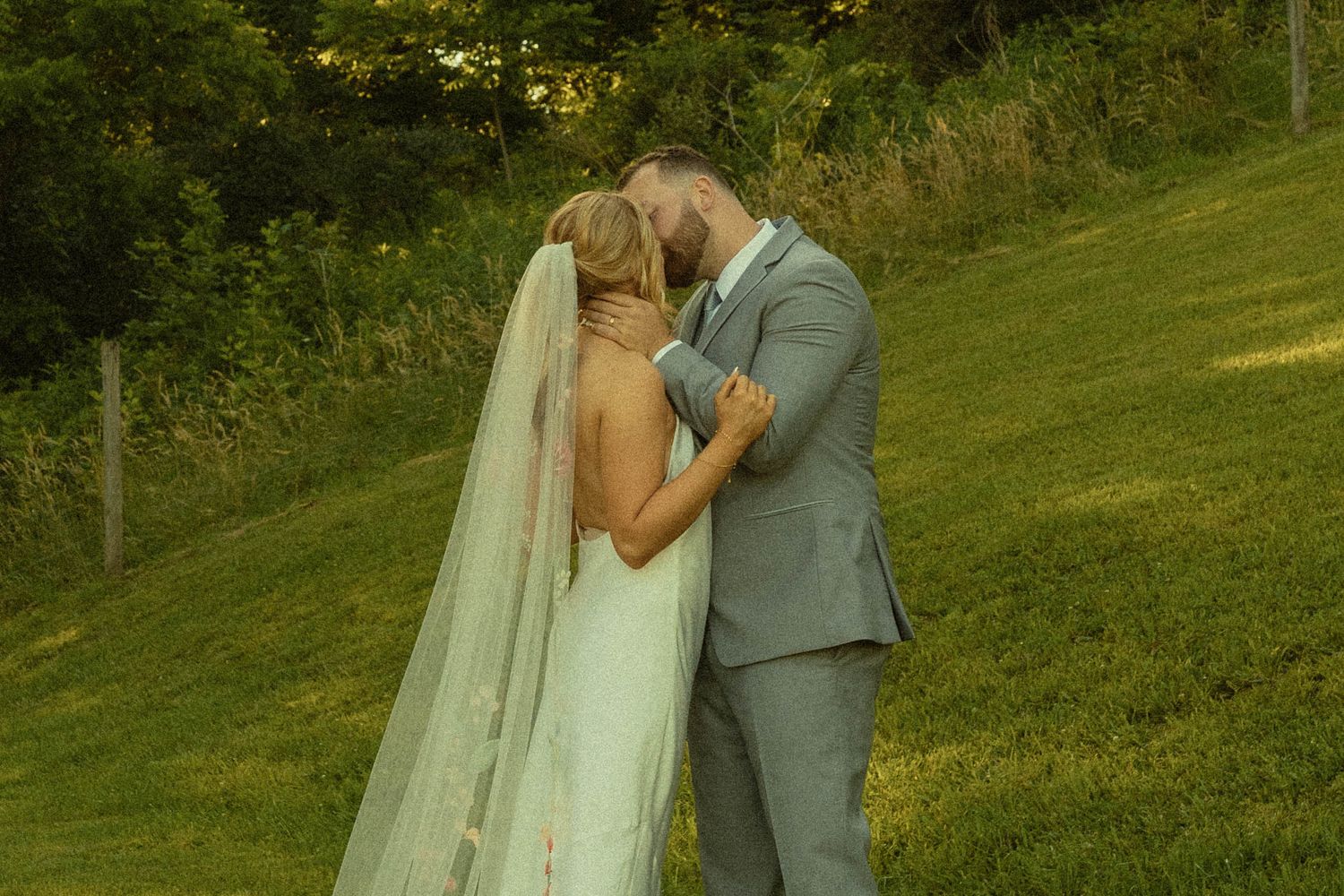 A romantic wedding couple shares an intimate moment on a sunlit grassy hillside.