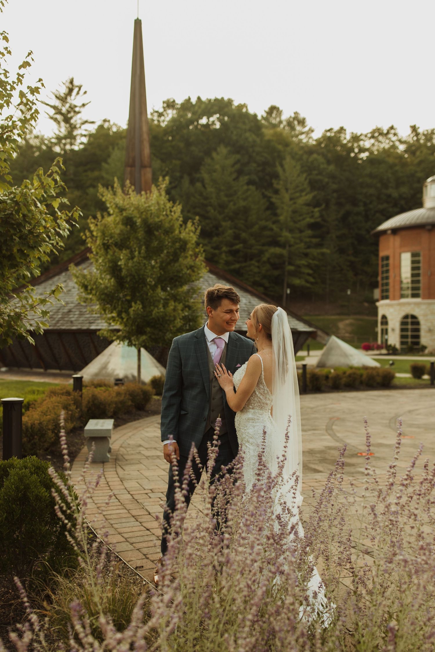 Wedding couple poses among purple flowers on brick pathway with church spire visible.