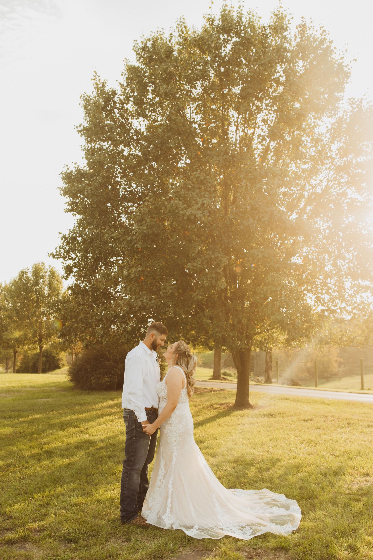 A romantic wedding portrait of a couple standing together in a sun-drenched field with tall trees.