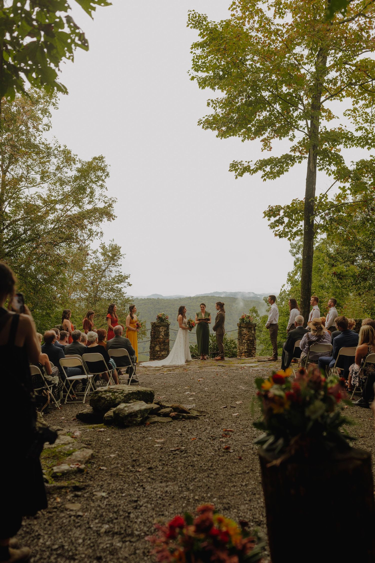 Outdoor forest wedding ceremony with guests seated on folding chairs and autumn floral decorations.