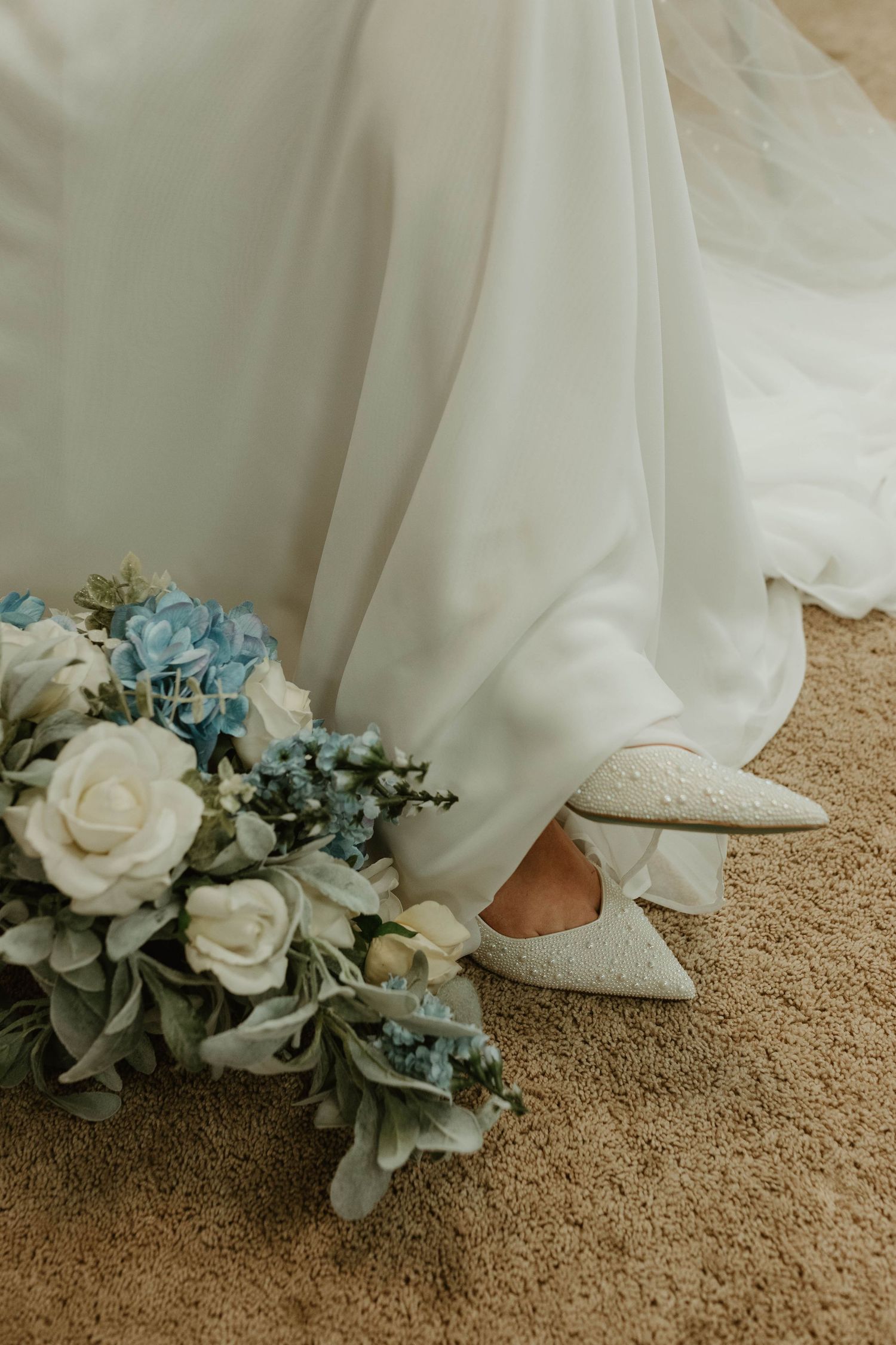 Wedding dress hem with blue and white floral bouquet and bridal shoes on carpet.