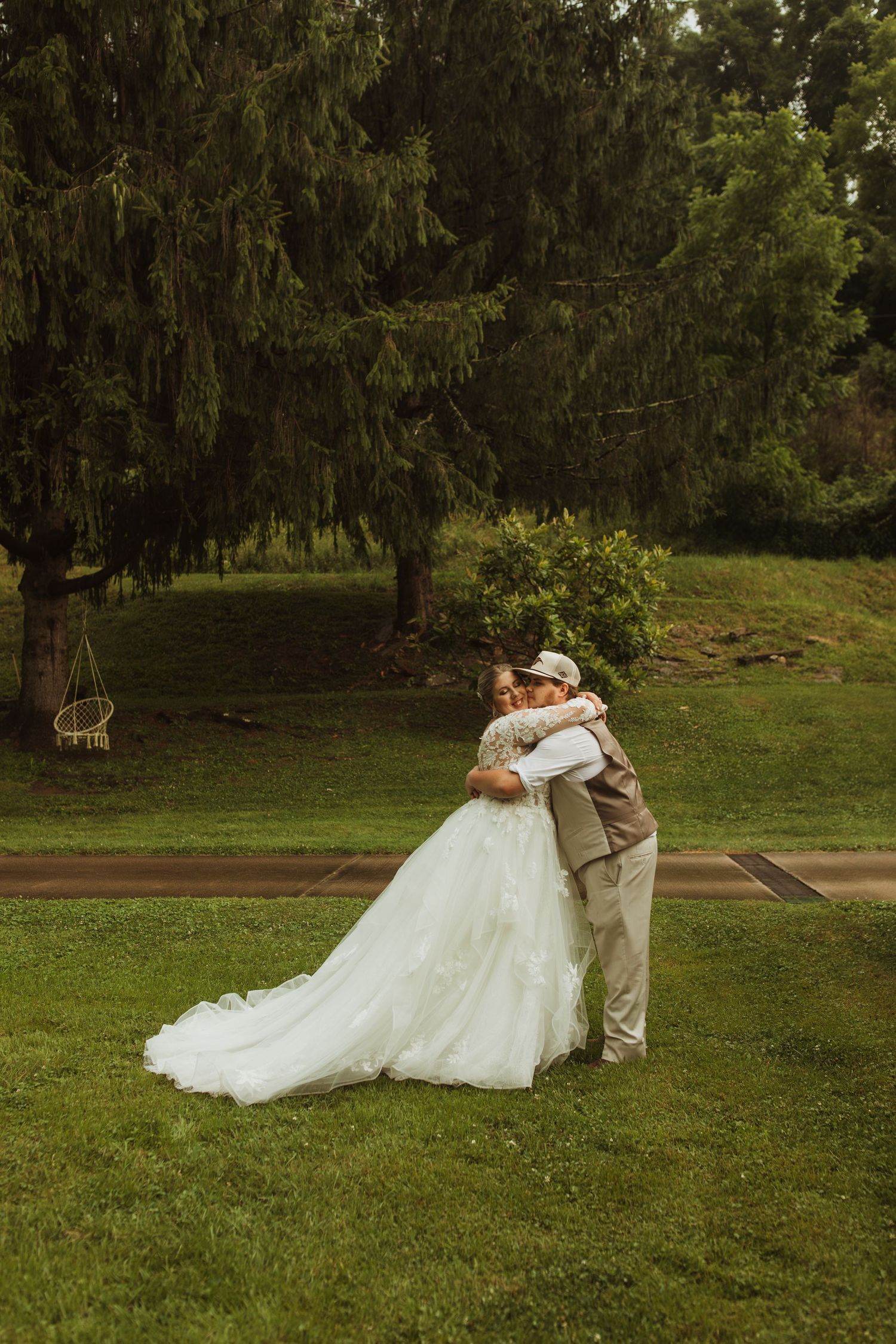 Couple embracing on grassy lawn beneath towering evergreen trees.