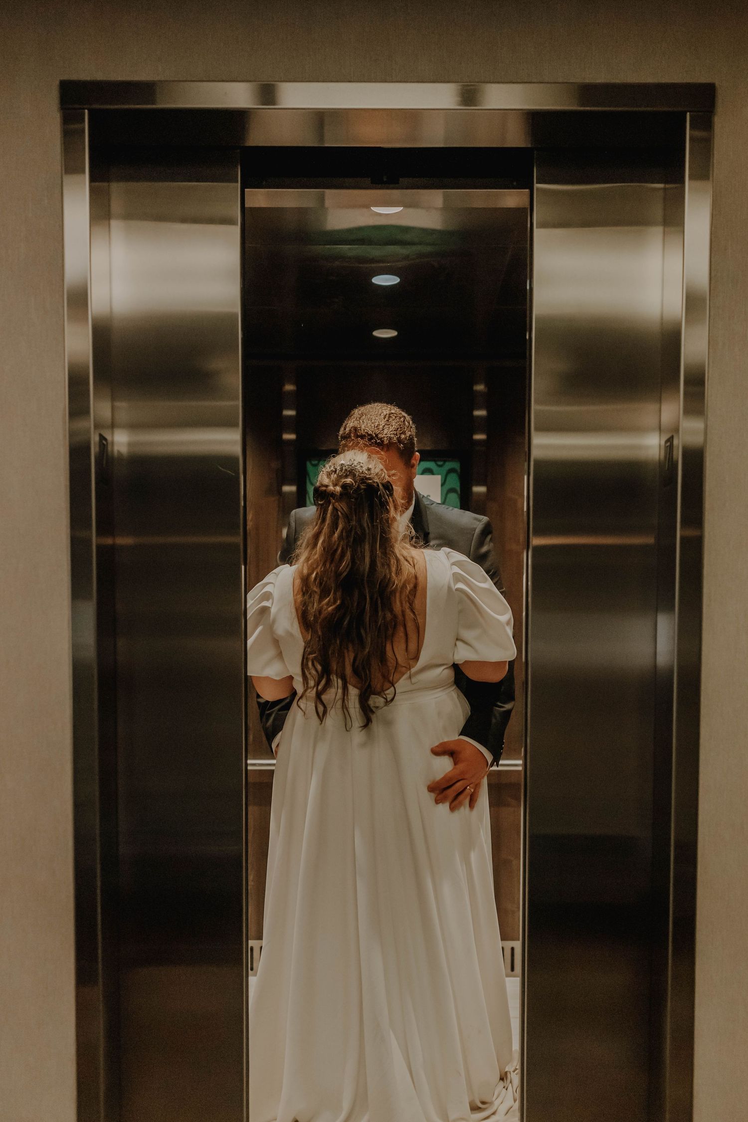 Elegant bride in a flowing white dress photographed from behind standing in a modern elevator.