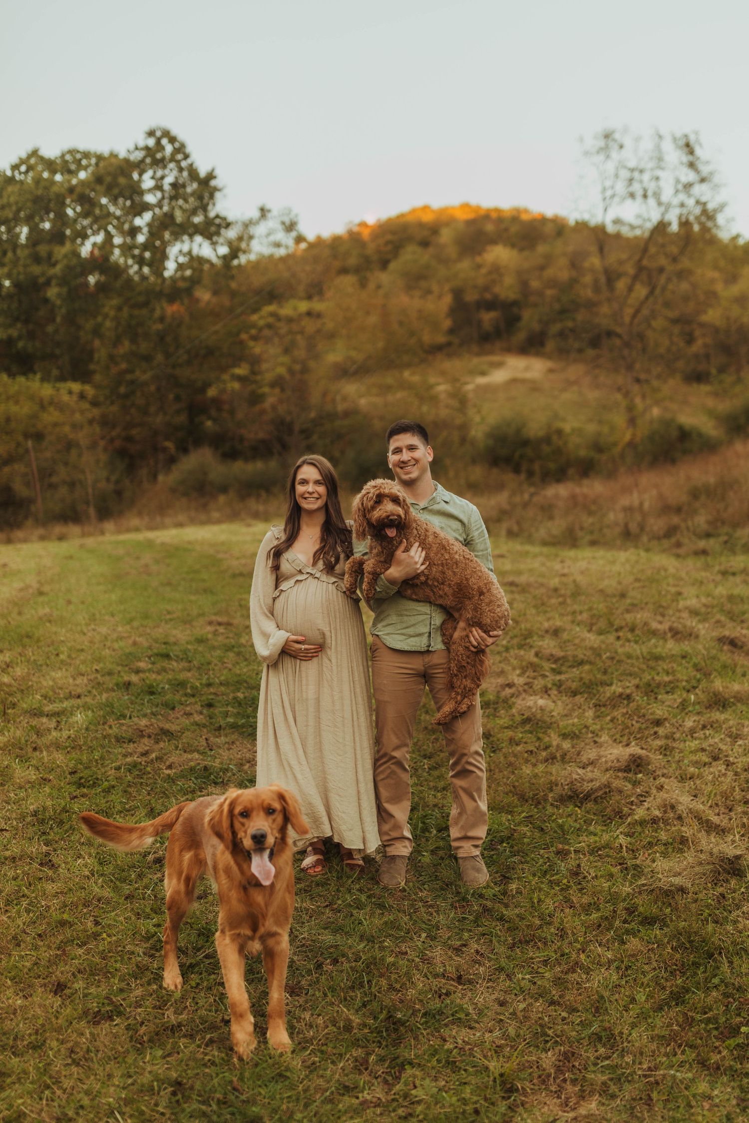 A couple poses with their two golden retriever dogs in a field during autumn.