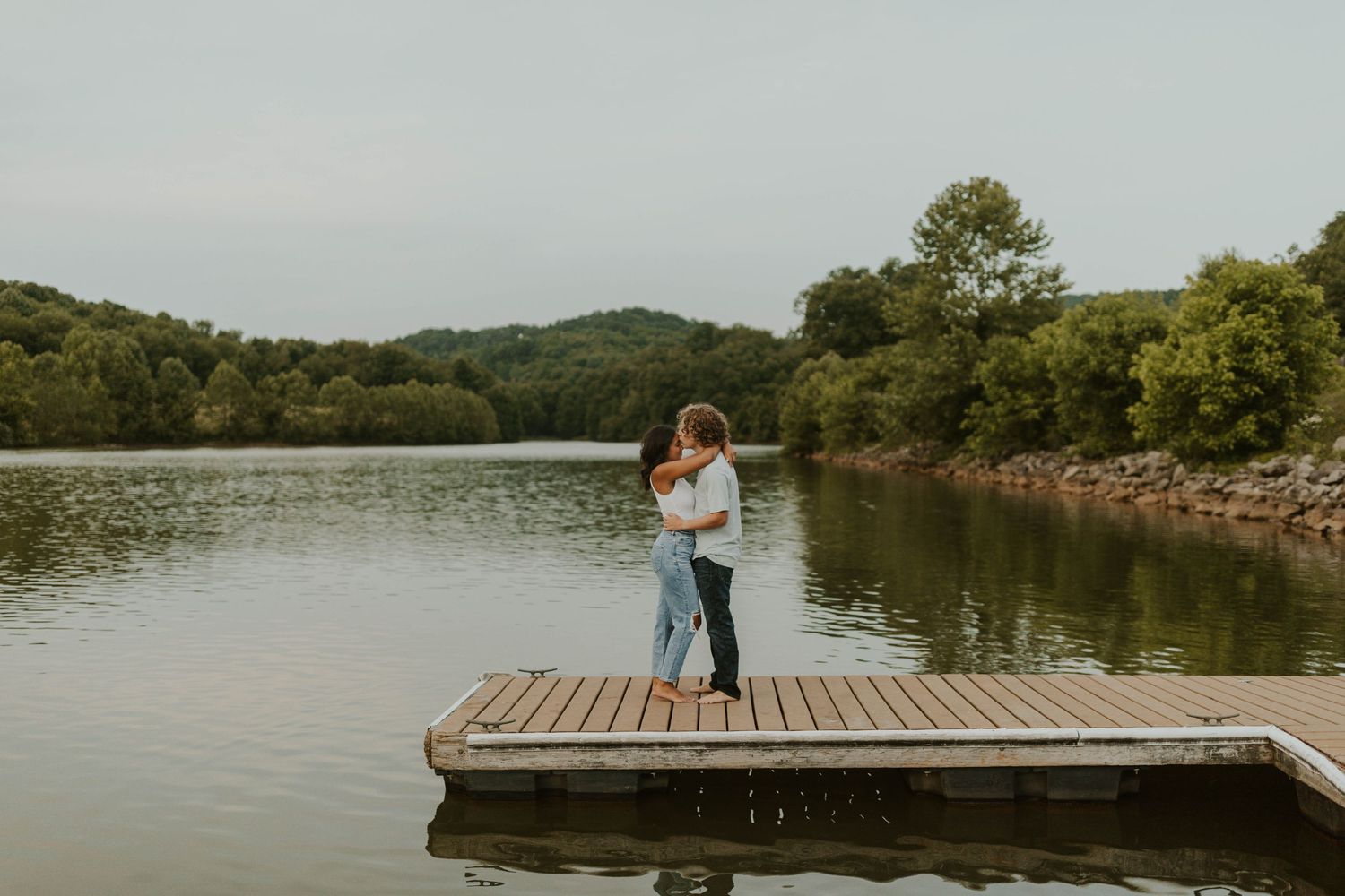 A couple embraces on a wooden dock overlooking a serene lake surrounded by hills.