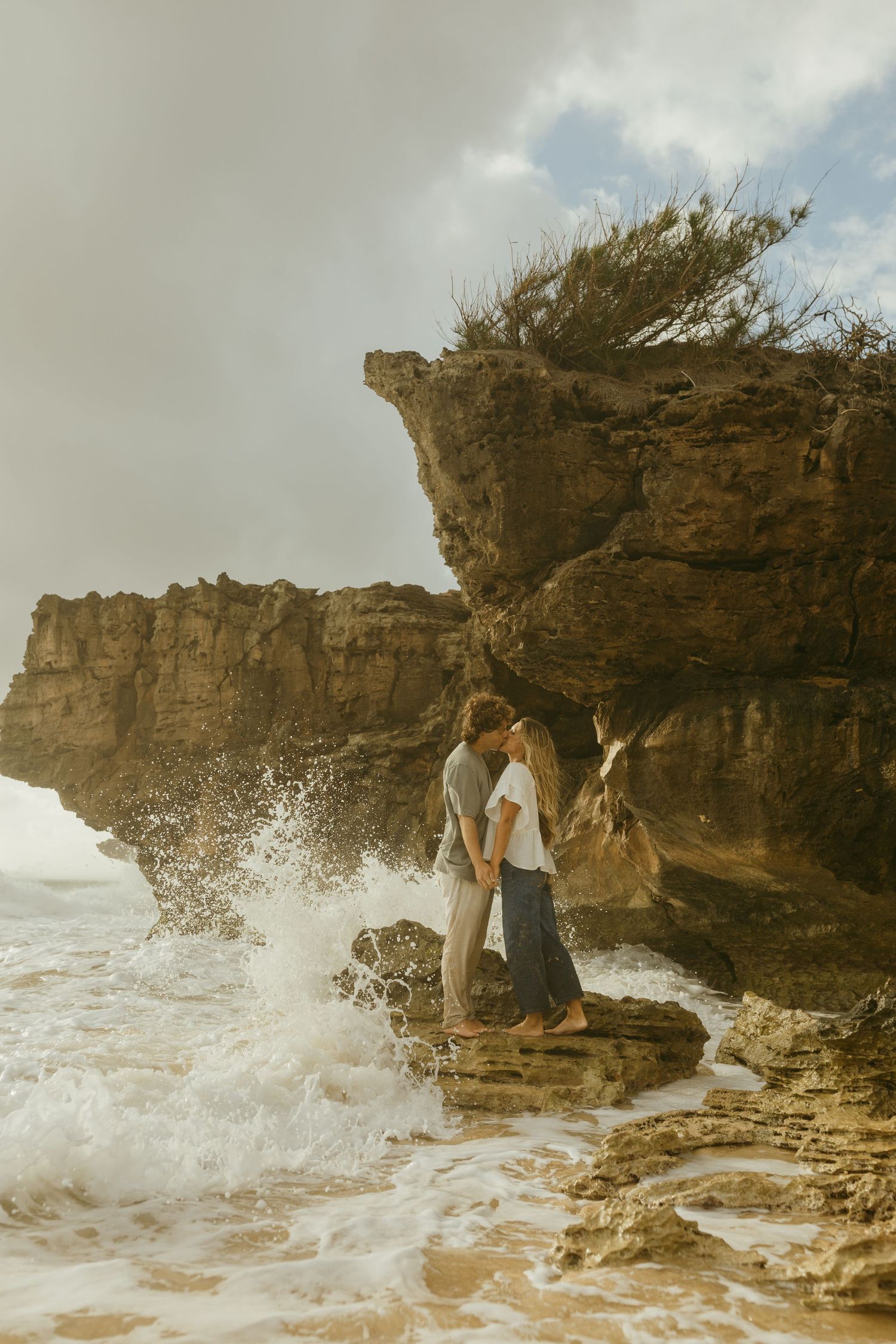 Couple embracing on rocky shore as waves crash against the cliffs.