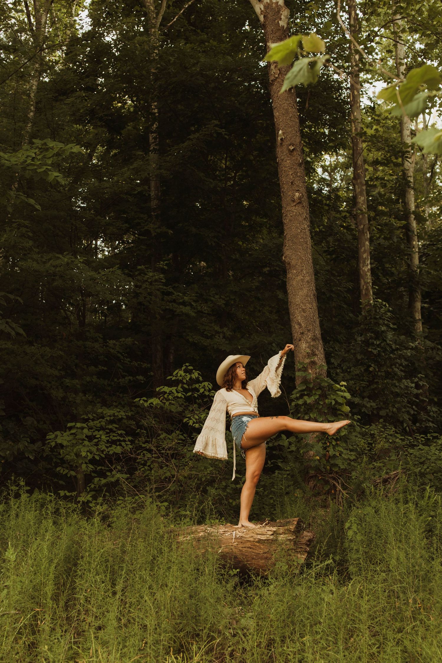 A dancer in a sun hat strikes an artistic pose on a tree stump in a wooded forest setting during golden hour.