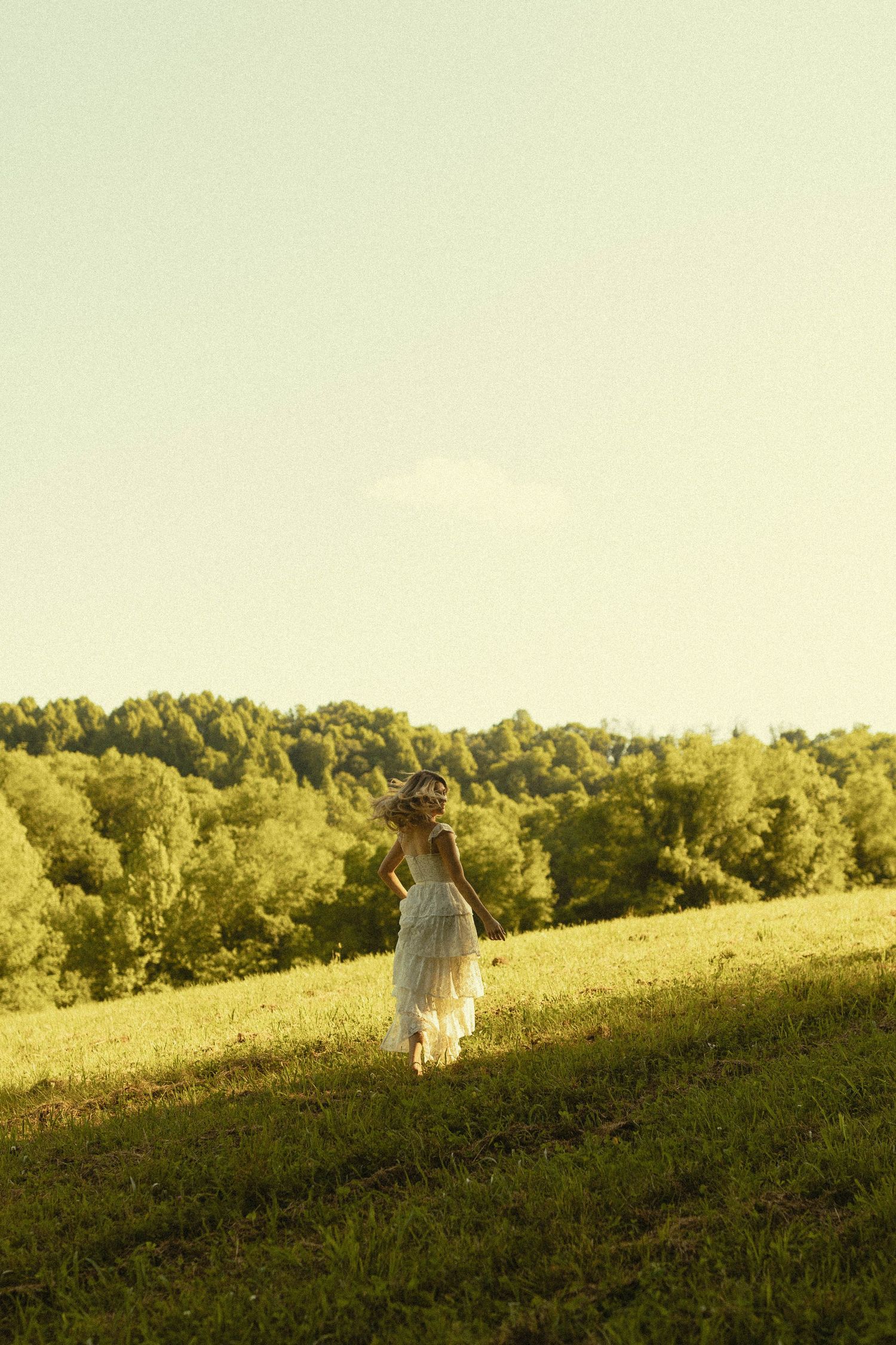 A dreamy silhouette in a flowing white dress stands on a hilltop at sunset.