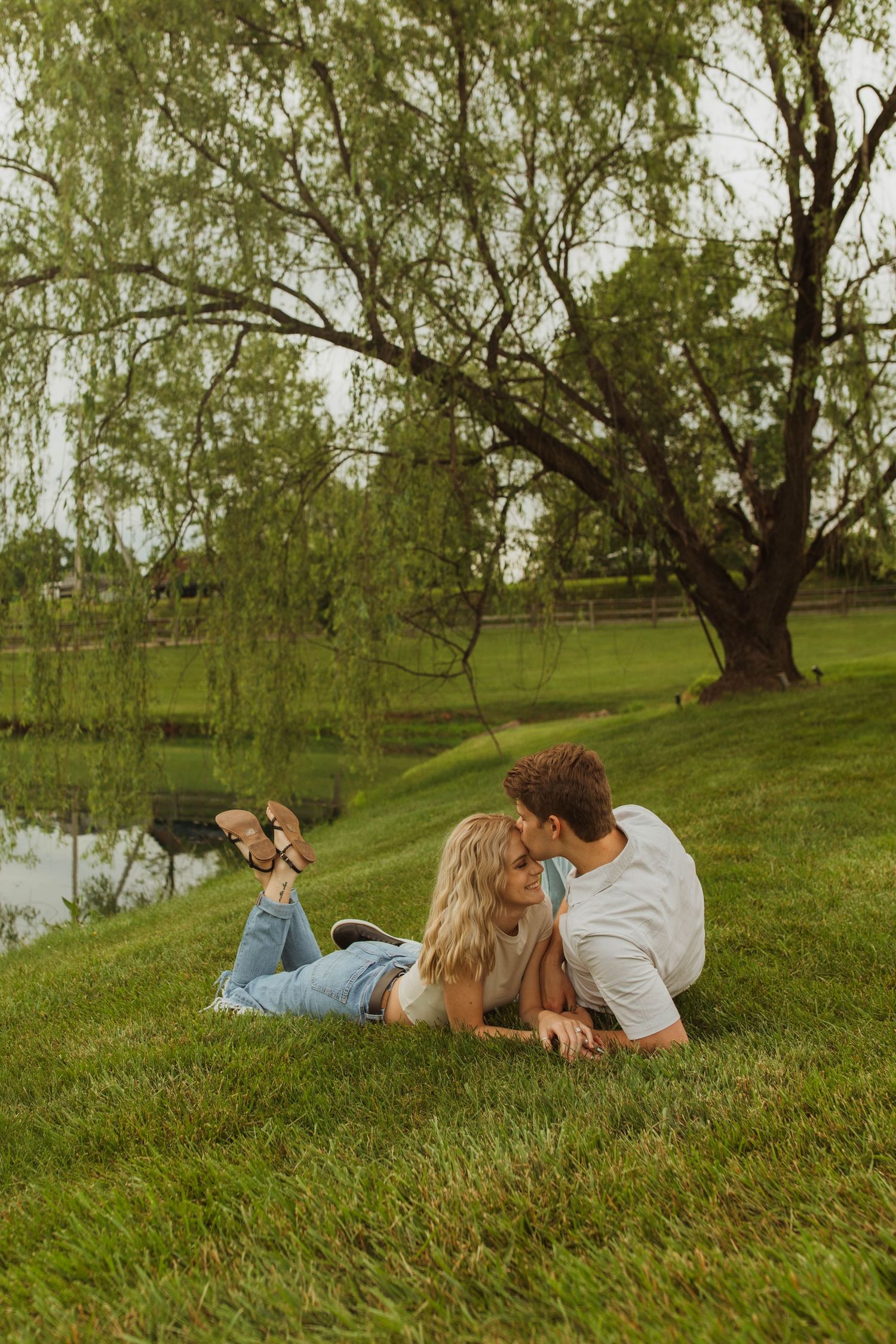 Couple relaxing together on grassy hill by pond under weeping willow tree.