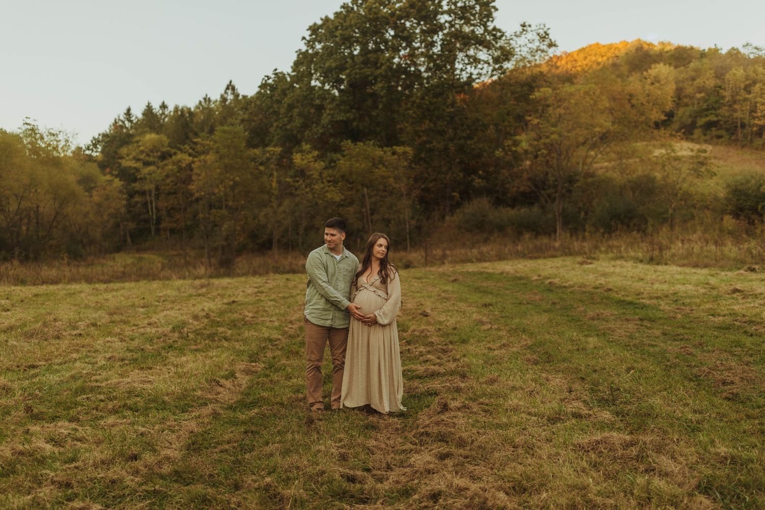 A couple stands together in a grassy field during sunset with golden hills in the background.
