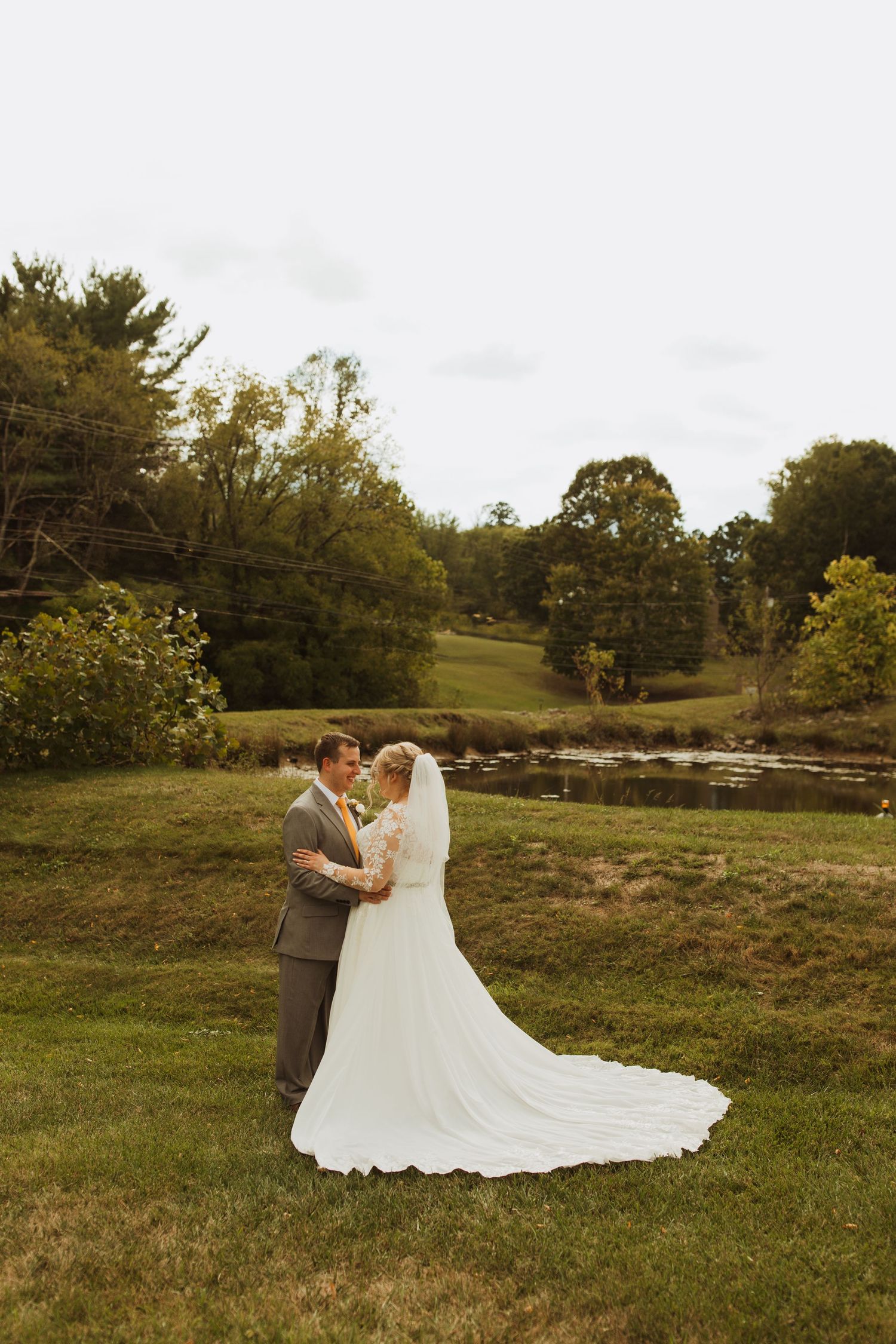 A bride and groom embrace in a scenic outdoor setting with a pond and autumn trees in the background.