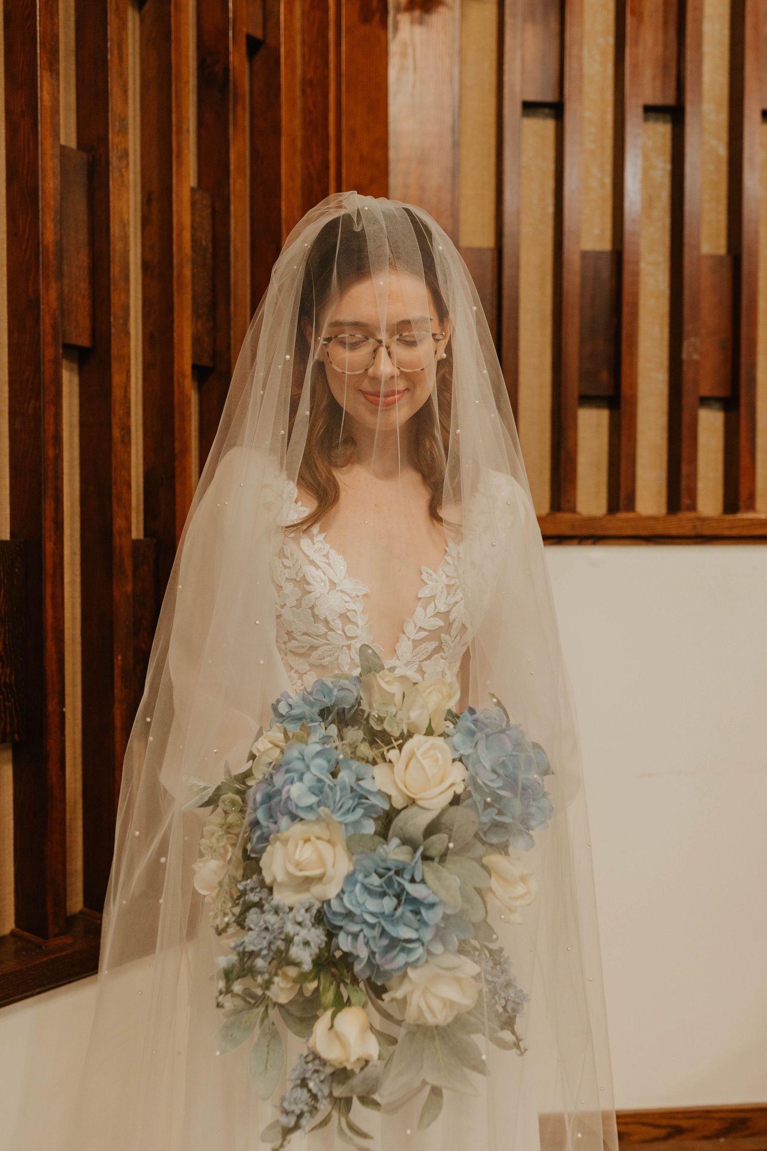 Bride holding blue and white bouquet wearing glasses and lace wedding dress.