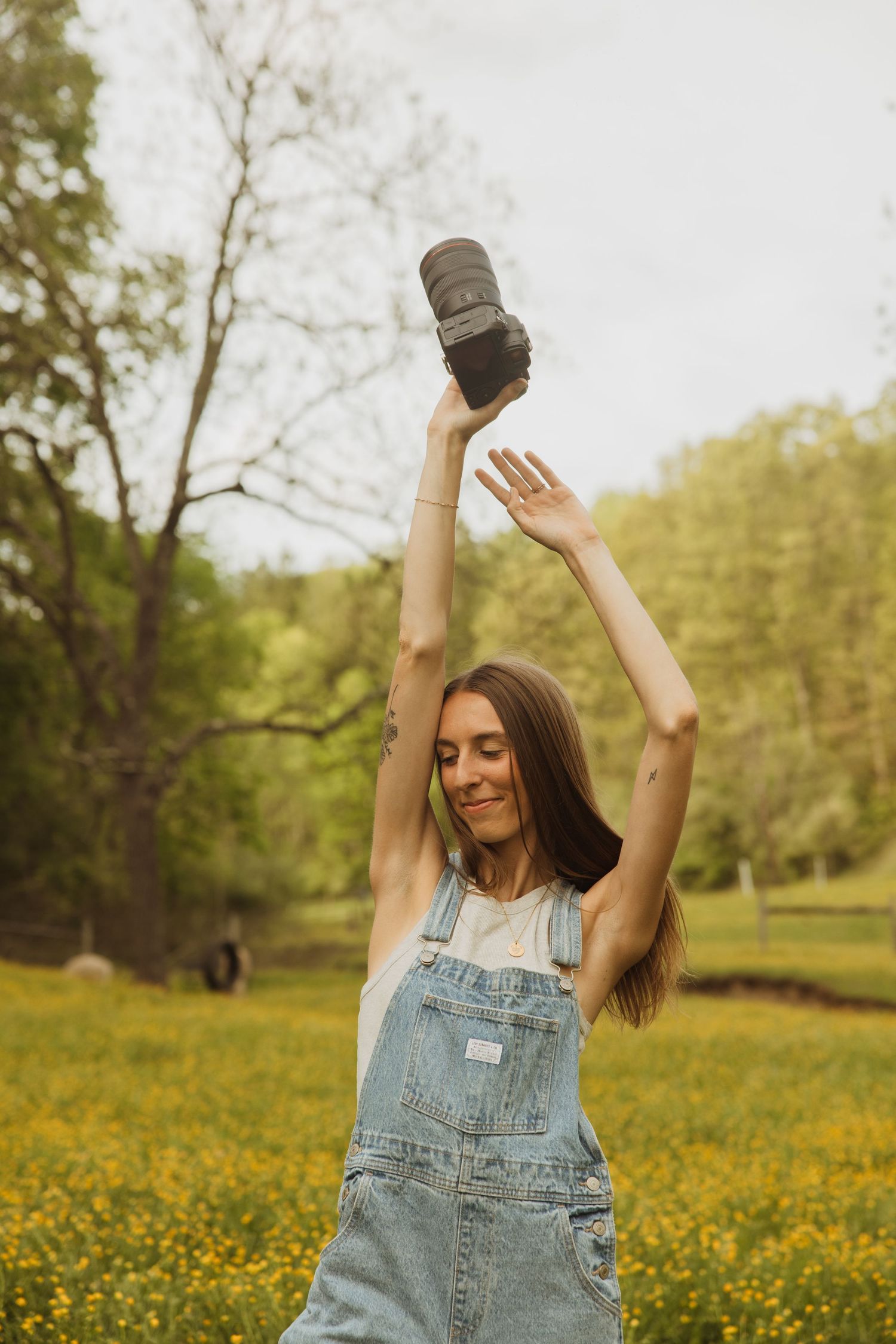 Person in denim overalls holds up a camera while dancing in a sunlit meadow filled with yellow wildflowers.