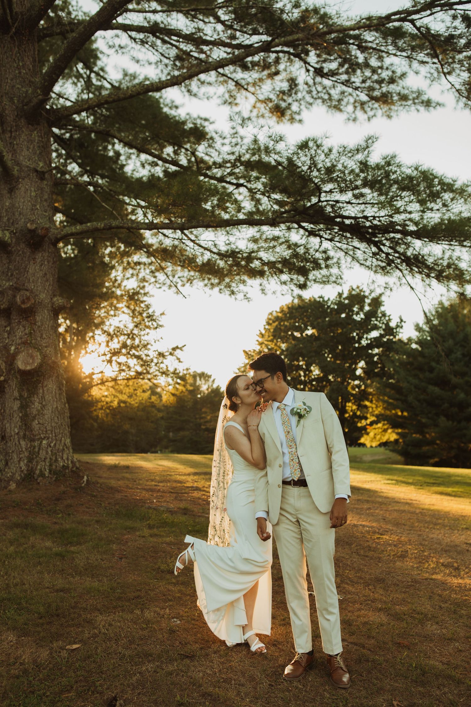 A romantic wedding couple embraces under a large pine tree at golden hour in a rustic outdoor setting.